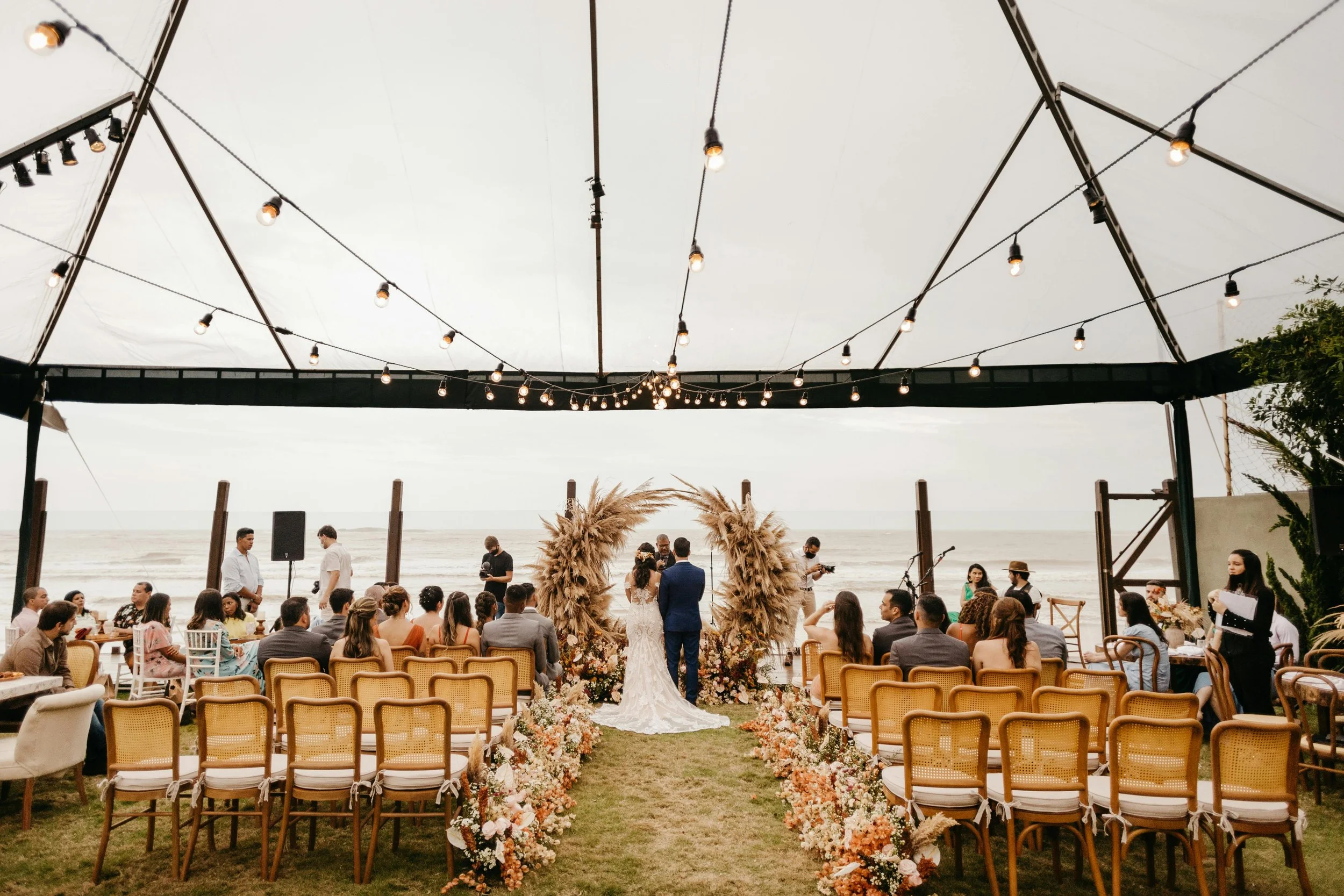 A beachside wedding ceremony with a bride and groom standing under a floral arch with guests seated on wooden chairs on either side, decorative flowers lining the aisle, and string lights hanging overhead under a large canopy.