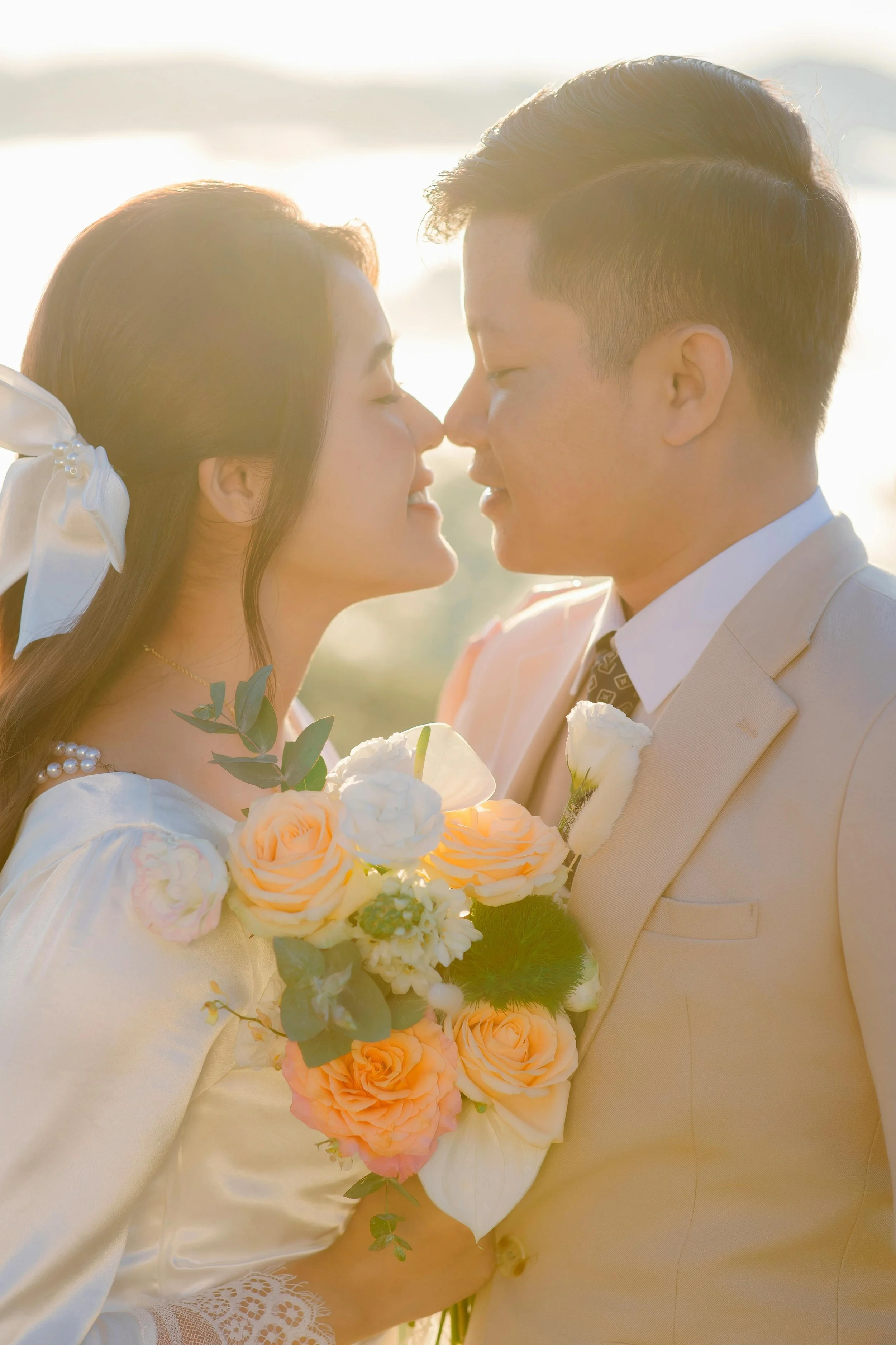 A couple on their wedding day, close together, smiling with noses touching, holding a bouquet of peach and white flowers, at sunset outdoor setting.