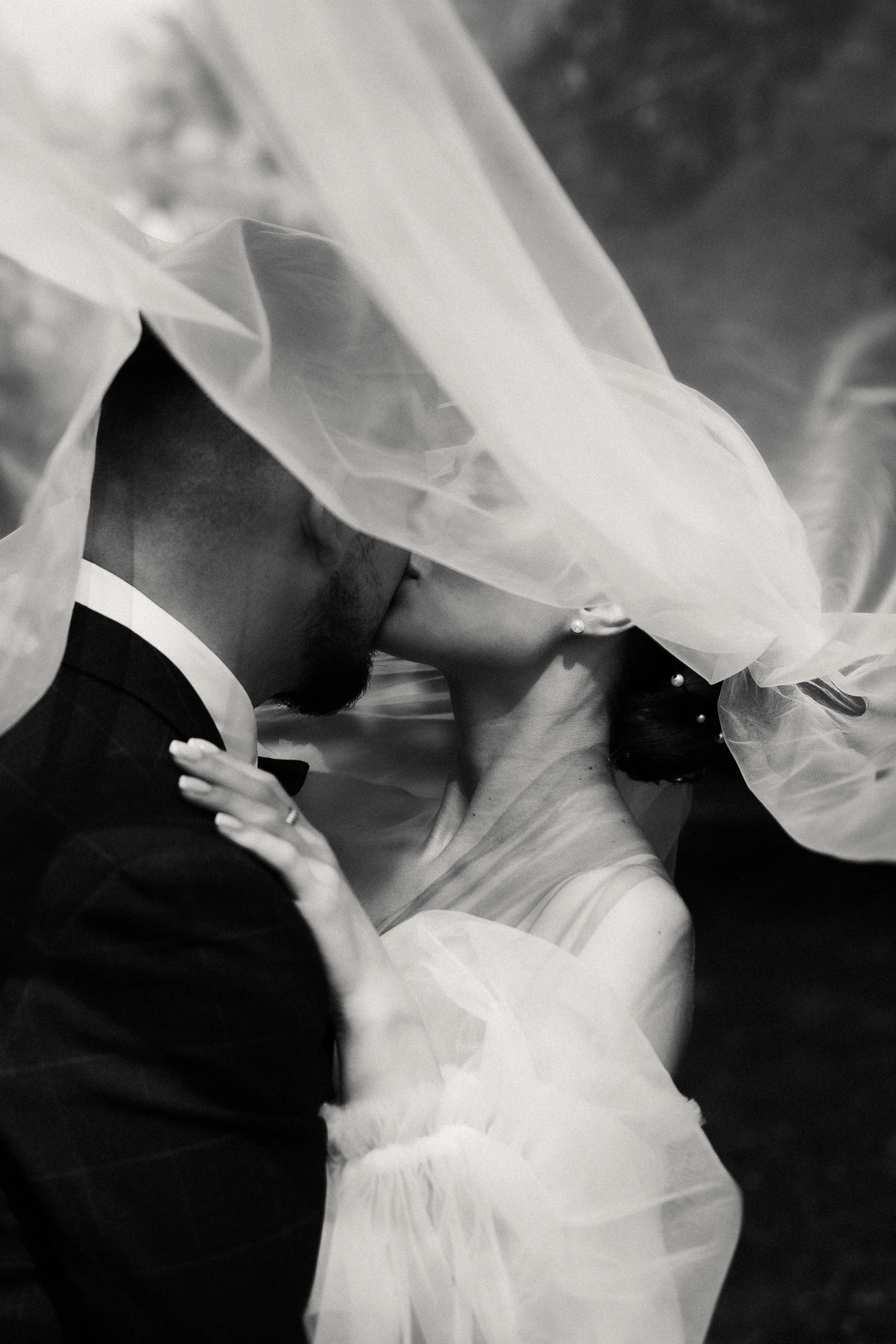 A black and white photo of a bride and groom sharing a kiss with the bride partially covered by a veil.