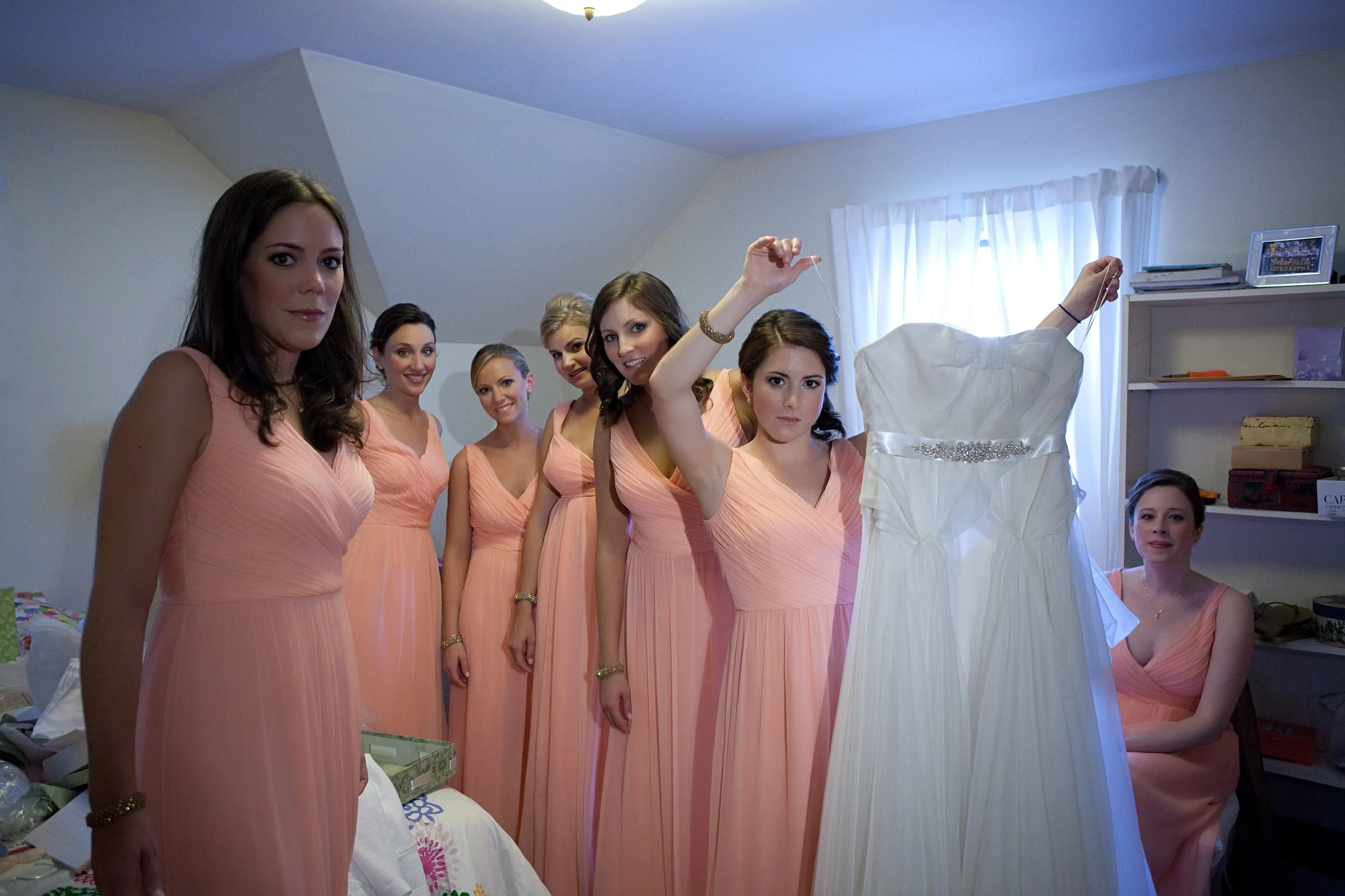 Six women in peach-colored dresses standing in a room, one woman holding a white wedding dress on a hanger, with a window and bookshelf in the background.