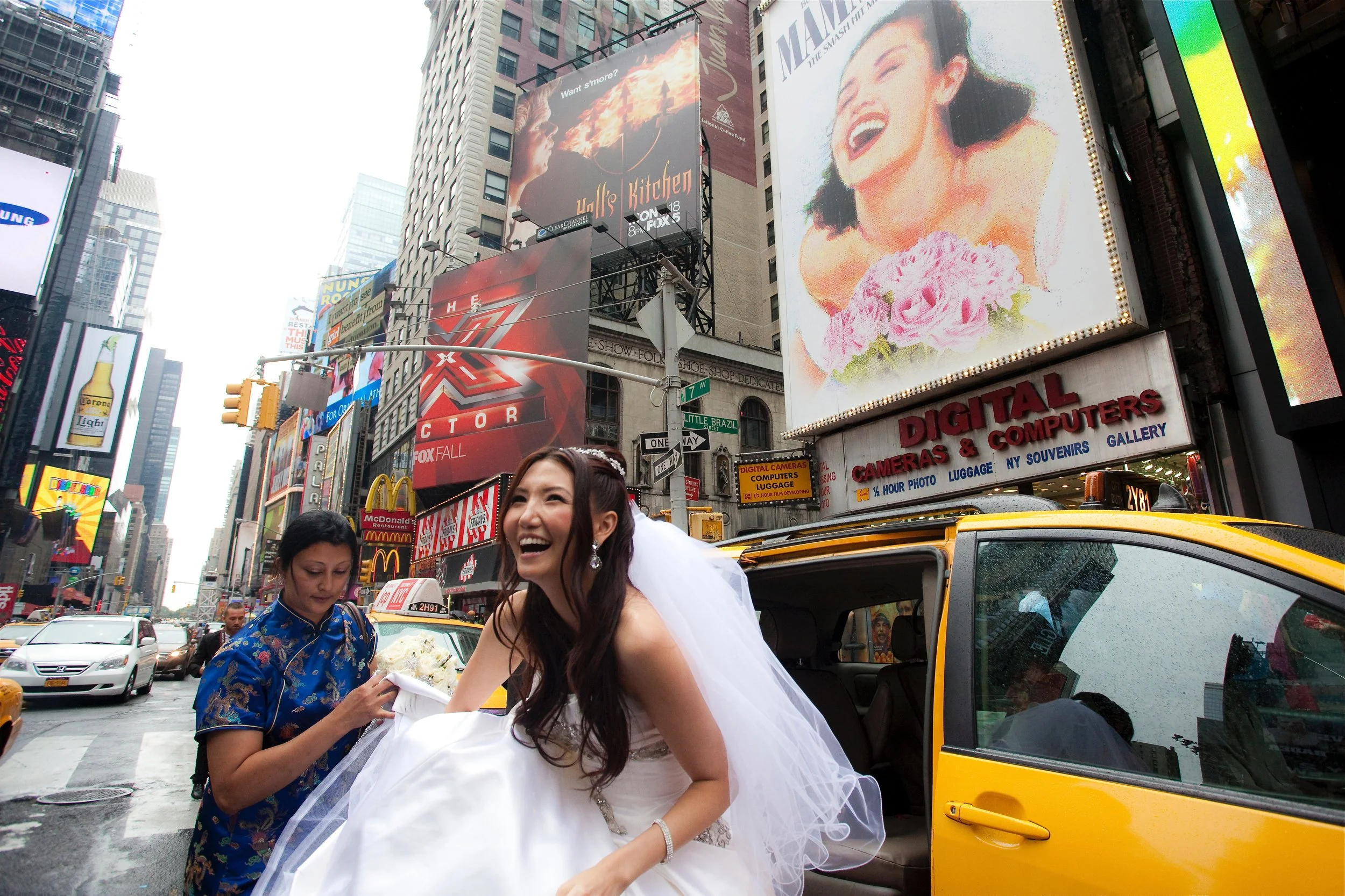 A joyful bride in a white wedding gown goes into a yellow taxi in Times Square, New York City, with a woman assisting her, surrounded by bright billboards and city traffic.