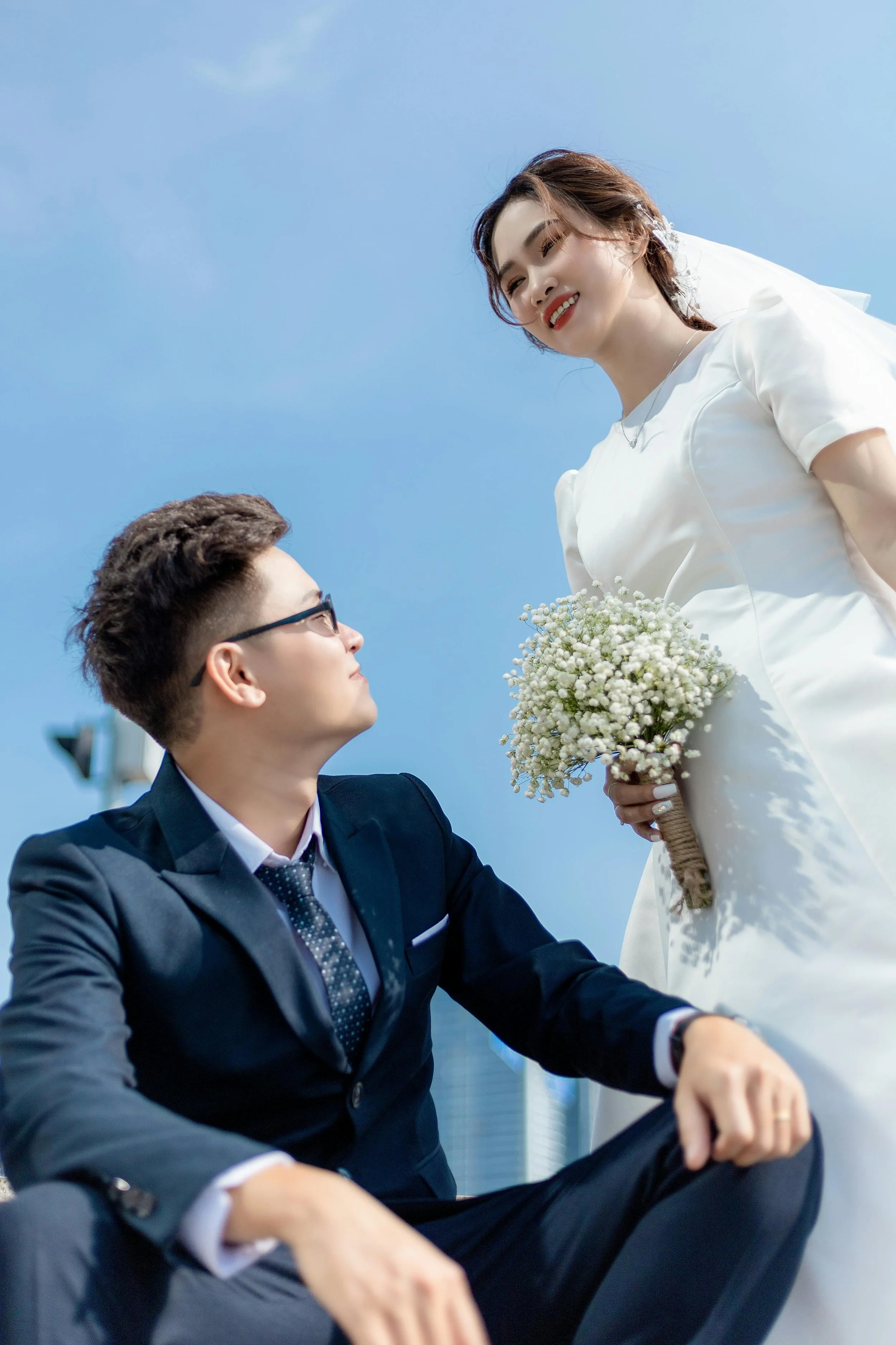A bride in white wedding dress holding a bouquet, looking down at a groom in a dark suit, sitting on the ground, with a blue sky background.