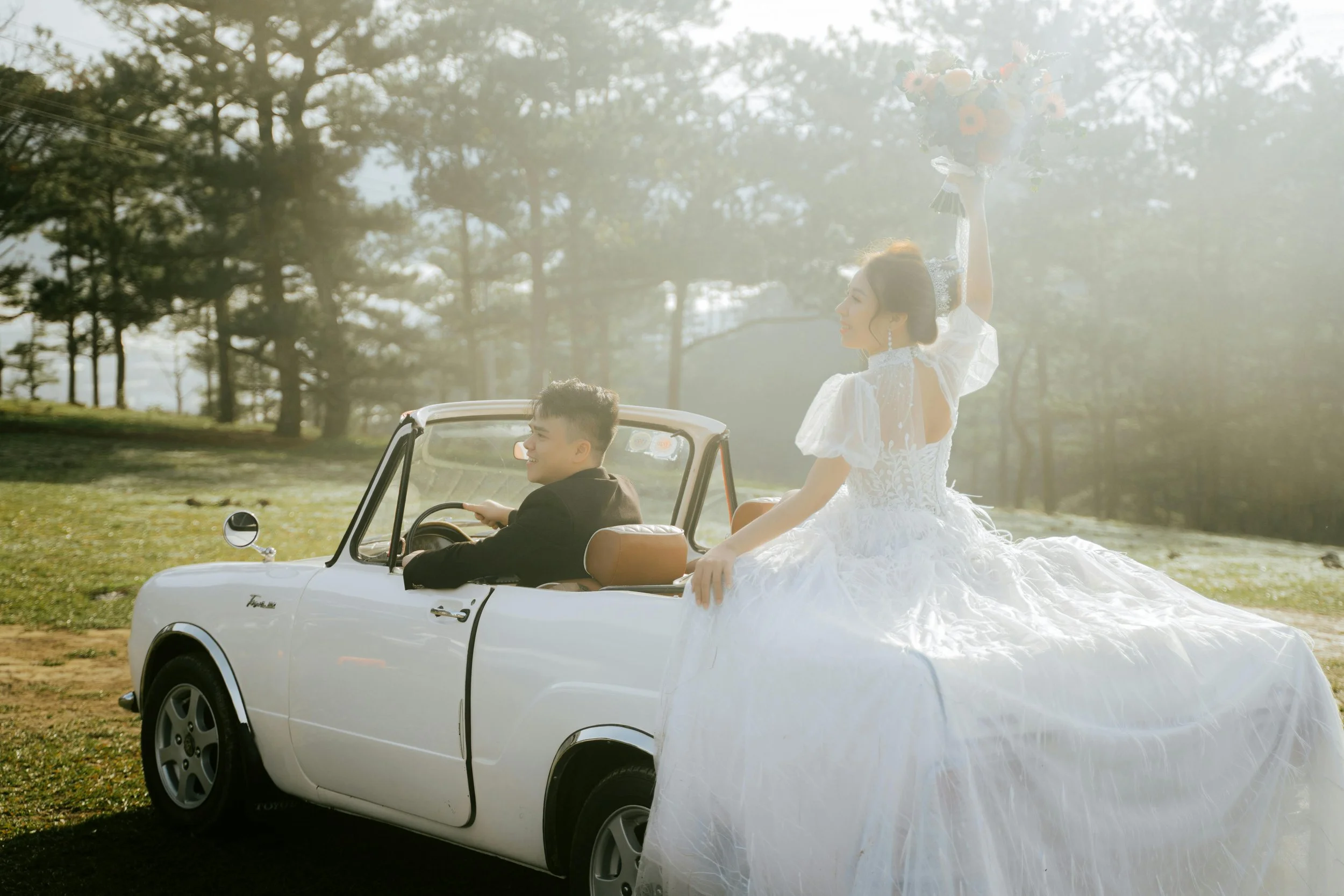A bride in a white wedding gown sitting on the back of a white vintage car with a male driver. The bride is holding a bouquet and has a joyful expression, with sunlight shining through the trees in the background.