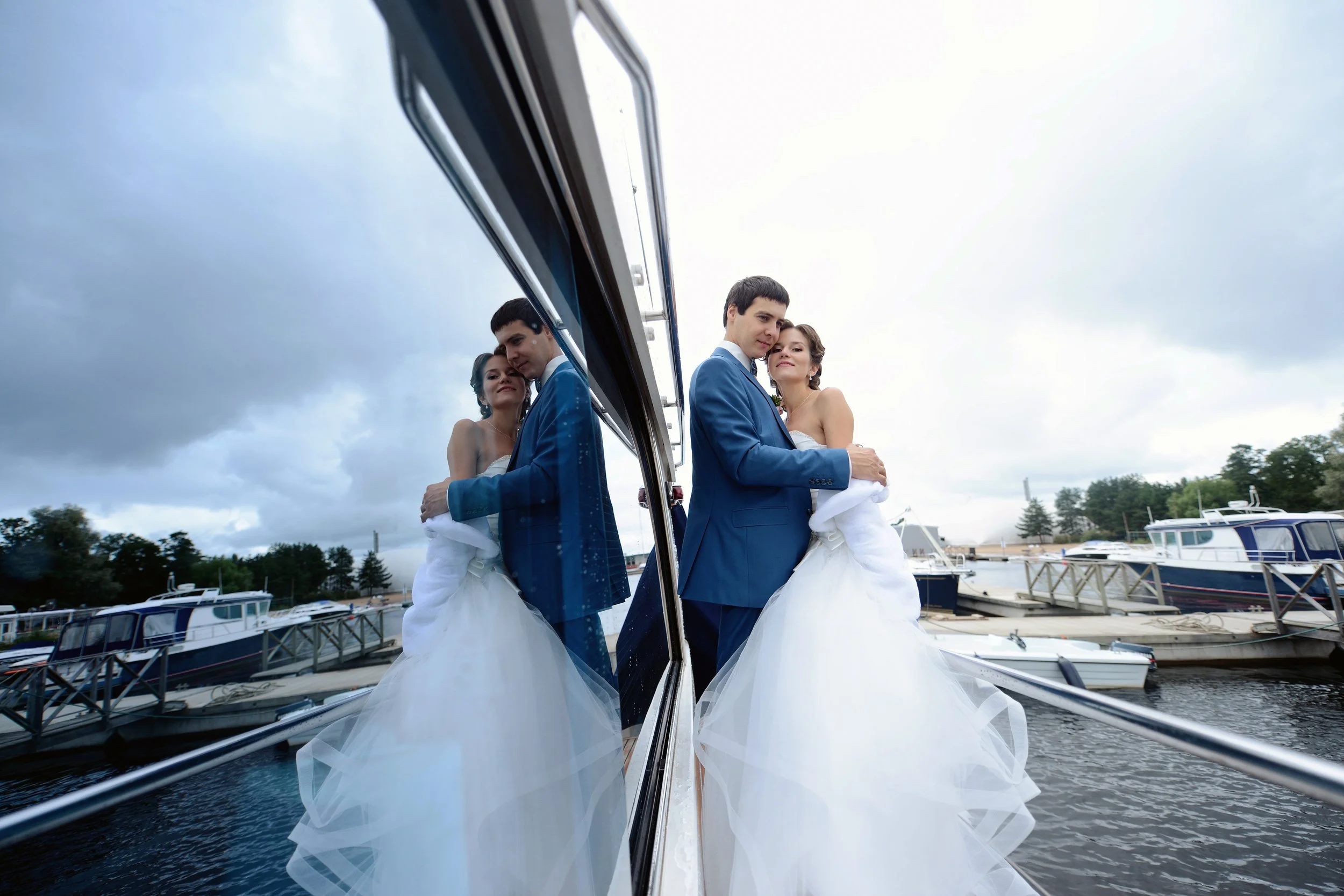 A bride and groom standing close together by a marina, reflected in a large mirror, with boats and cloudy sky in the background.