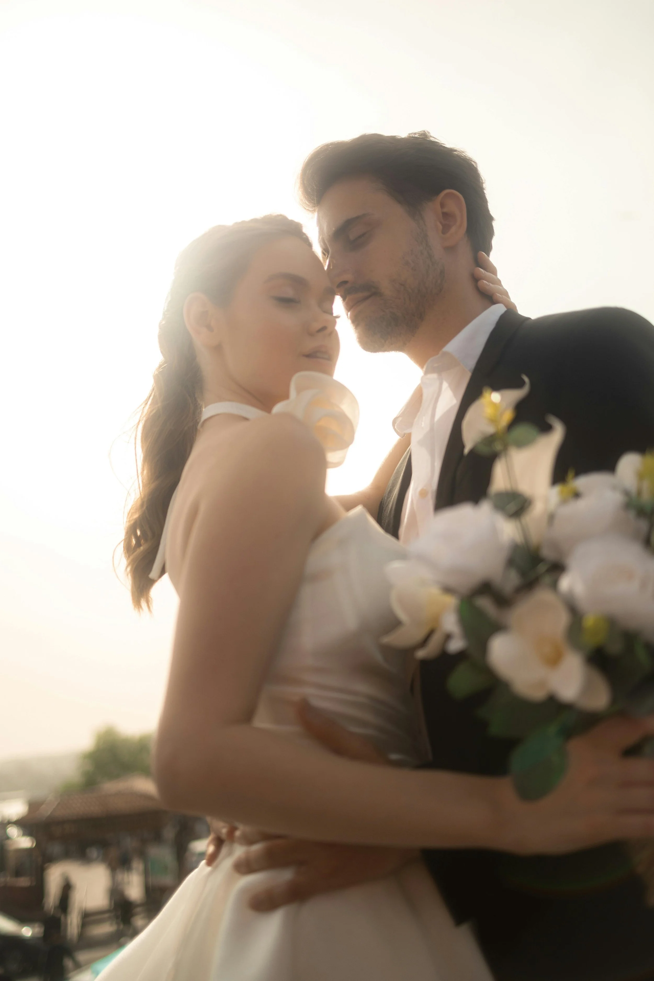 A bride and groom embrace outdoors at sunset, the bride holding a bouquet of white flowers, both with their eyes closed and foreheads touching.