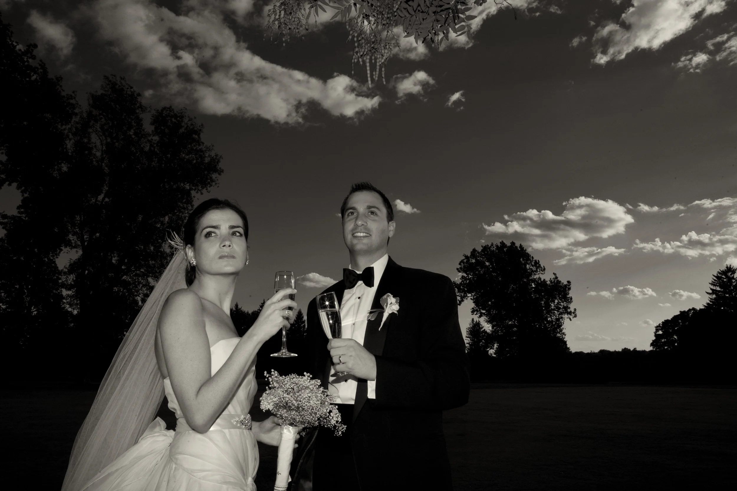 Black and white photo of a bride and groom holding glasses of champagne outdoors, with trees and cloudy sky in the background.