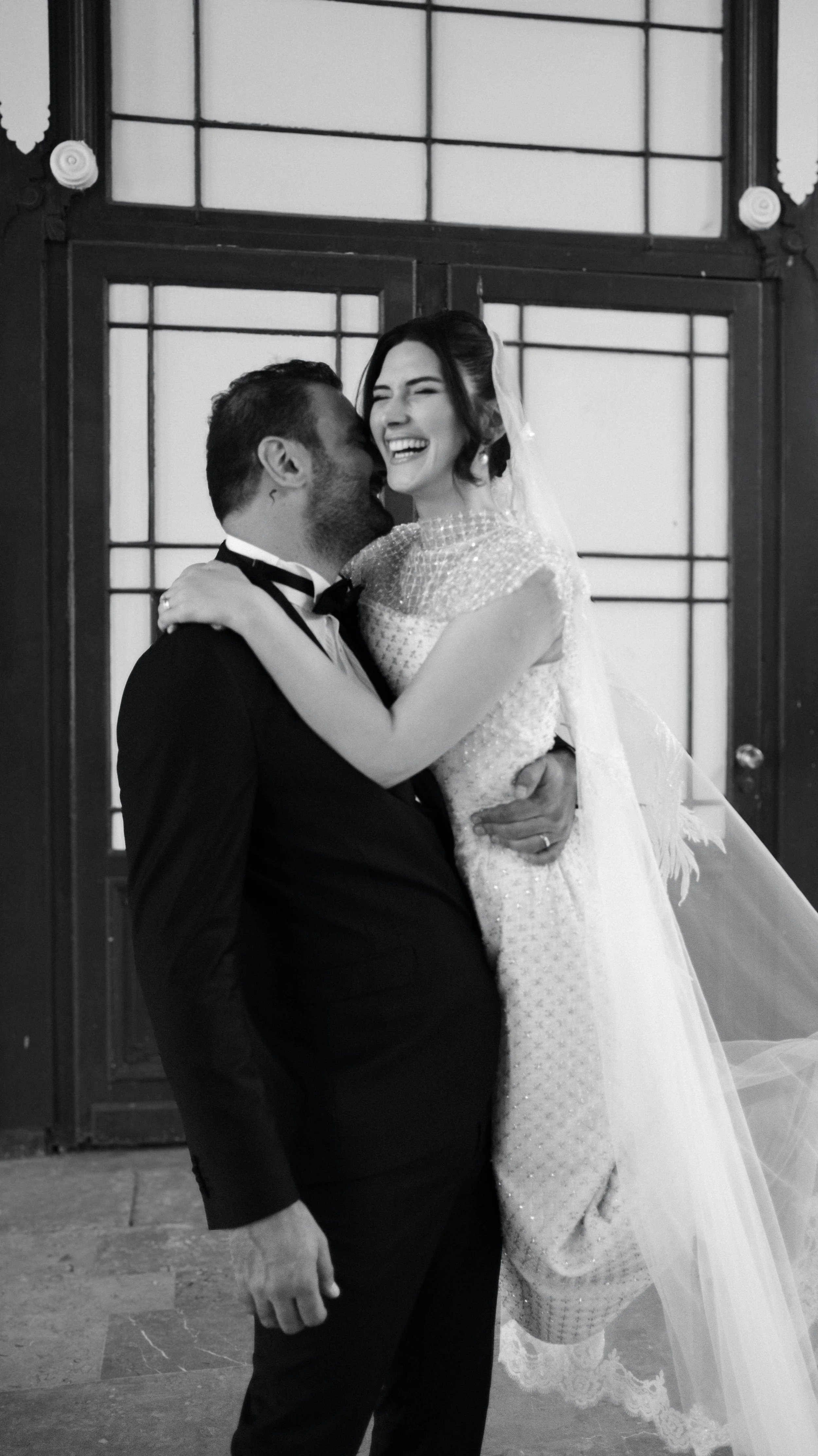 A happy bride and groom sharing a joyful moment, with the bride in a wedding dress and veil, and the groom in a tuxedo, standing in front of a large, decorative door.