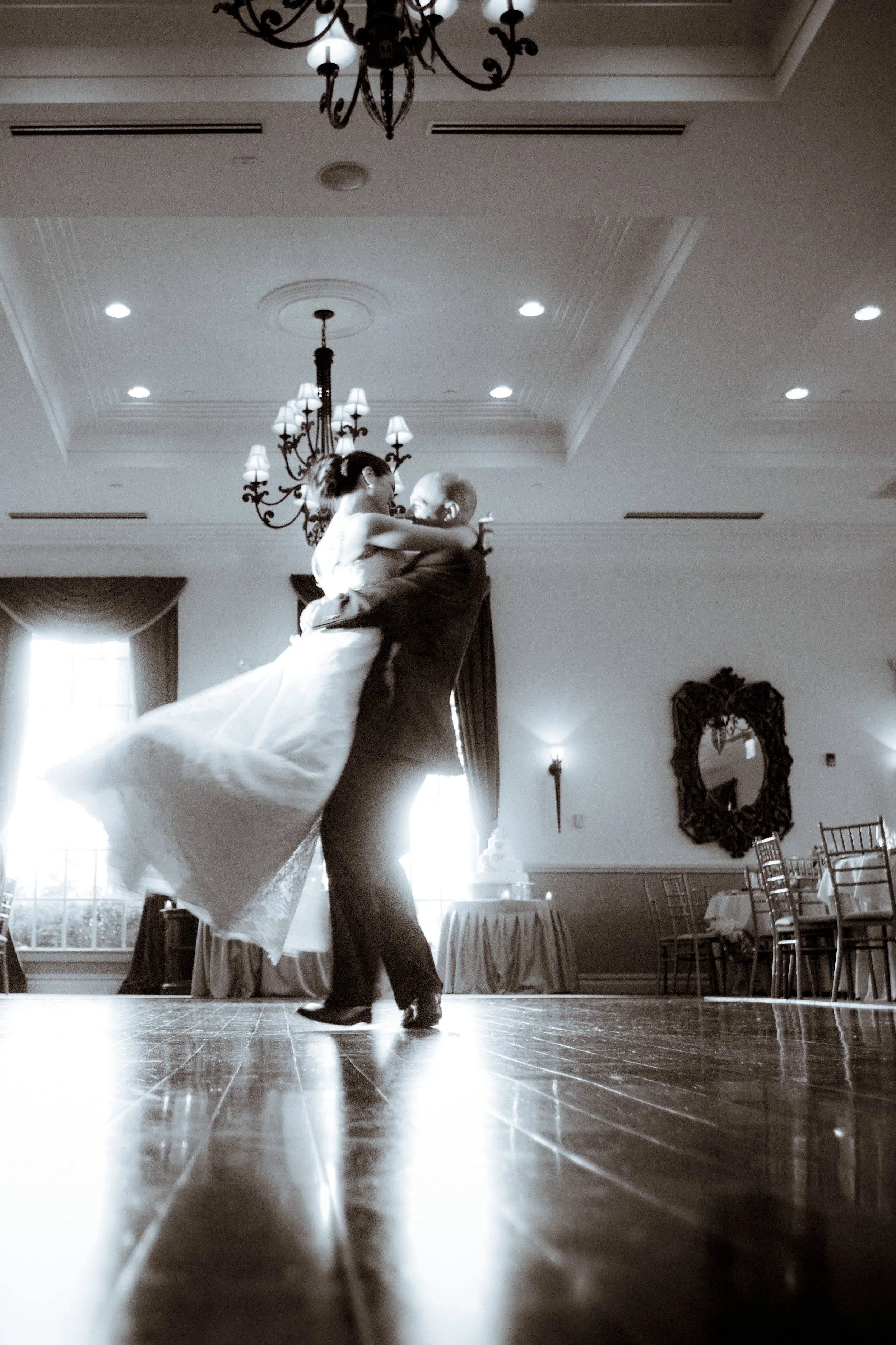 A bride and groom dancing at a wedding reception in an elegant indoor venue with chandeliers and large windows.