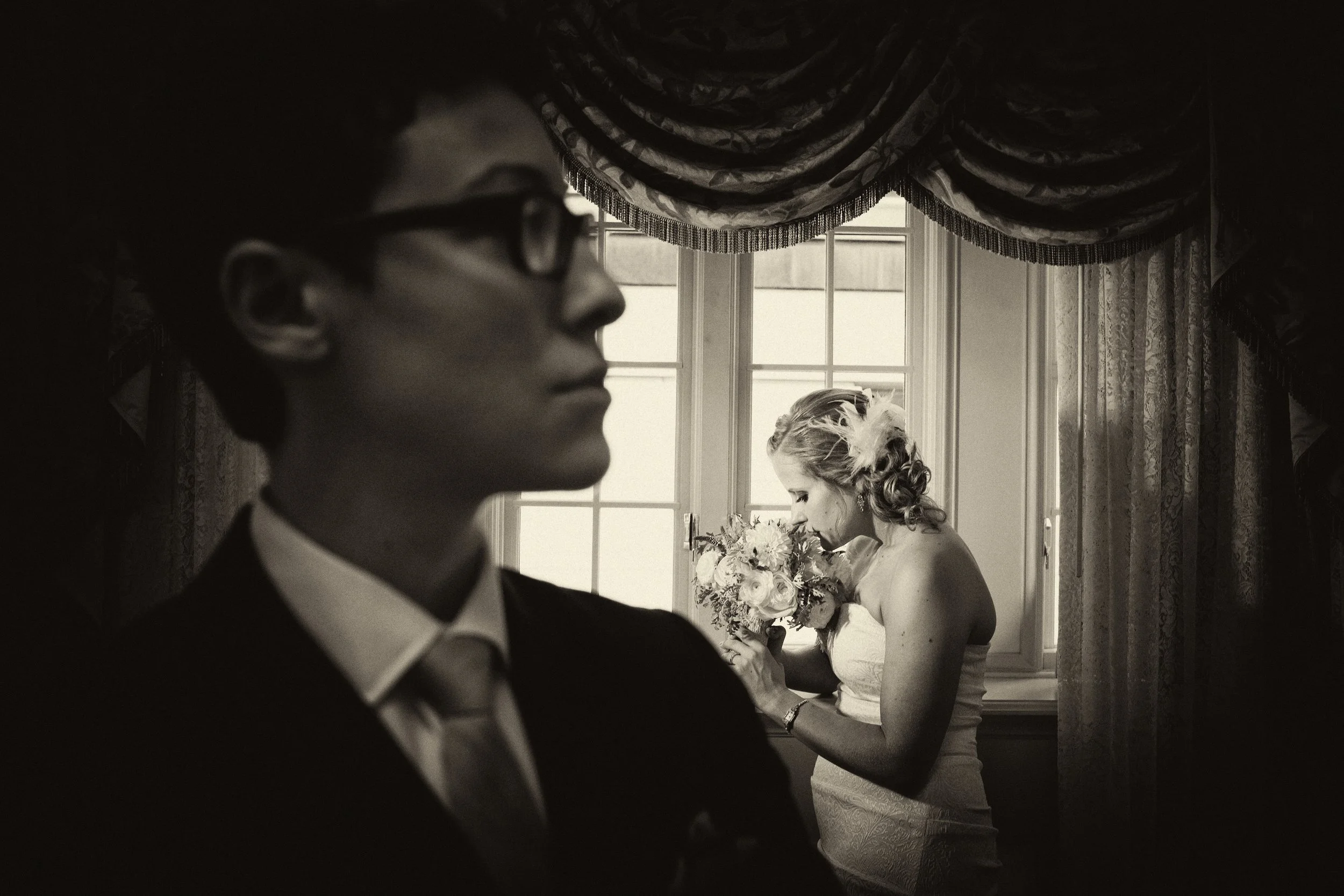 A black and white photo of a groom in glasses in the foreground and a bride holding a bouquet near a window in the background, both dressed in wedding attire.
