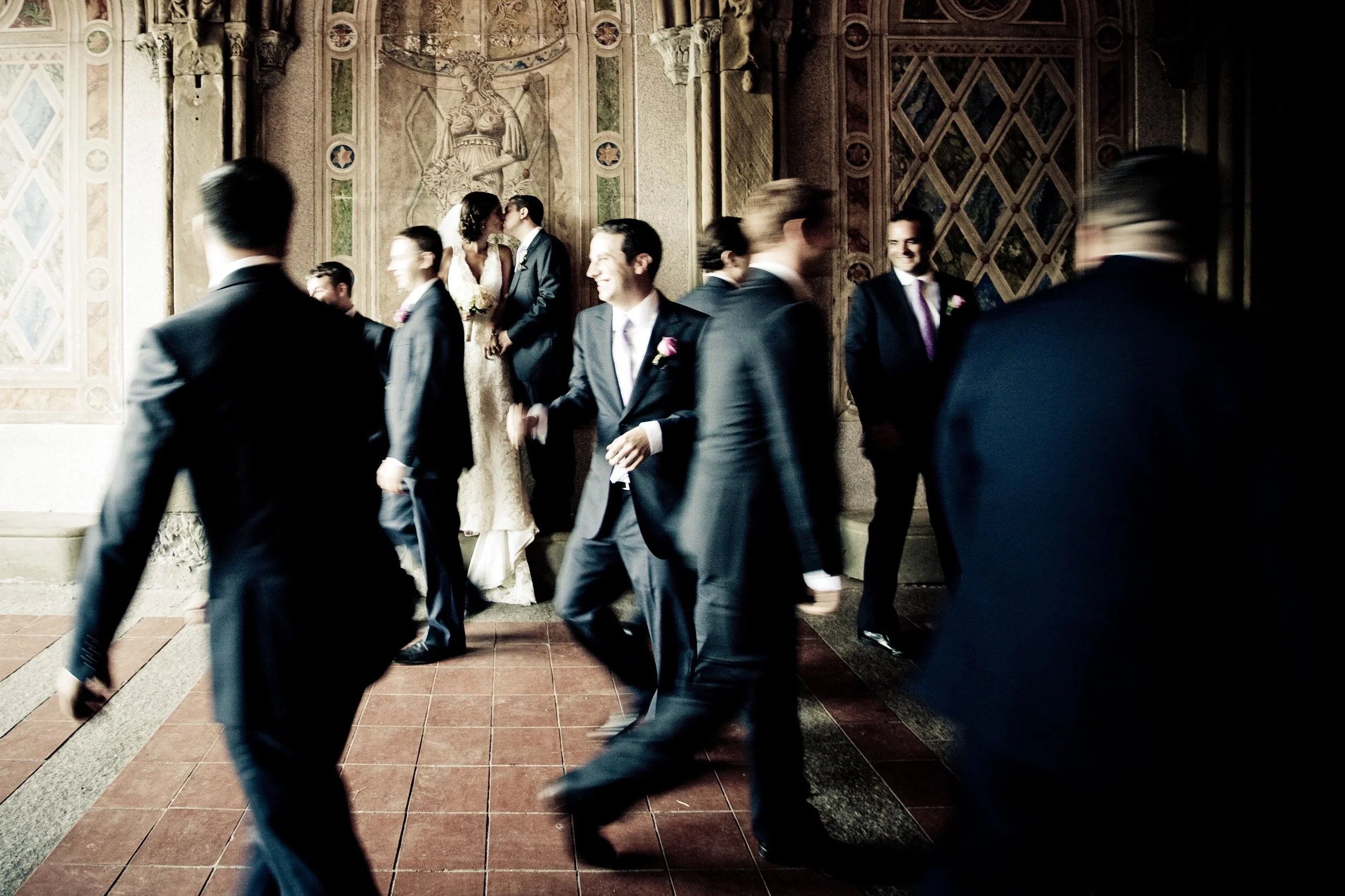 Wedding reception with the bride and groom kissing on a decorated wall, surrounded by guests in formal attire, some walking and others standing, in an elegant venue with ornate wall art.