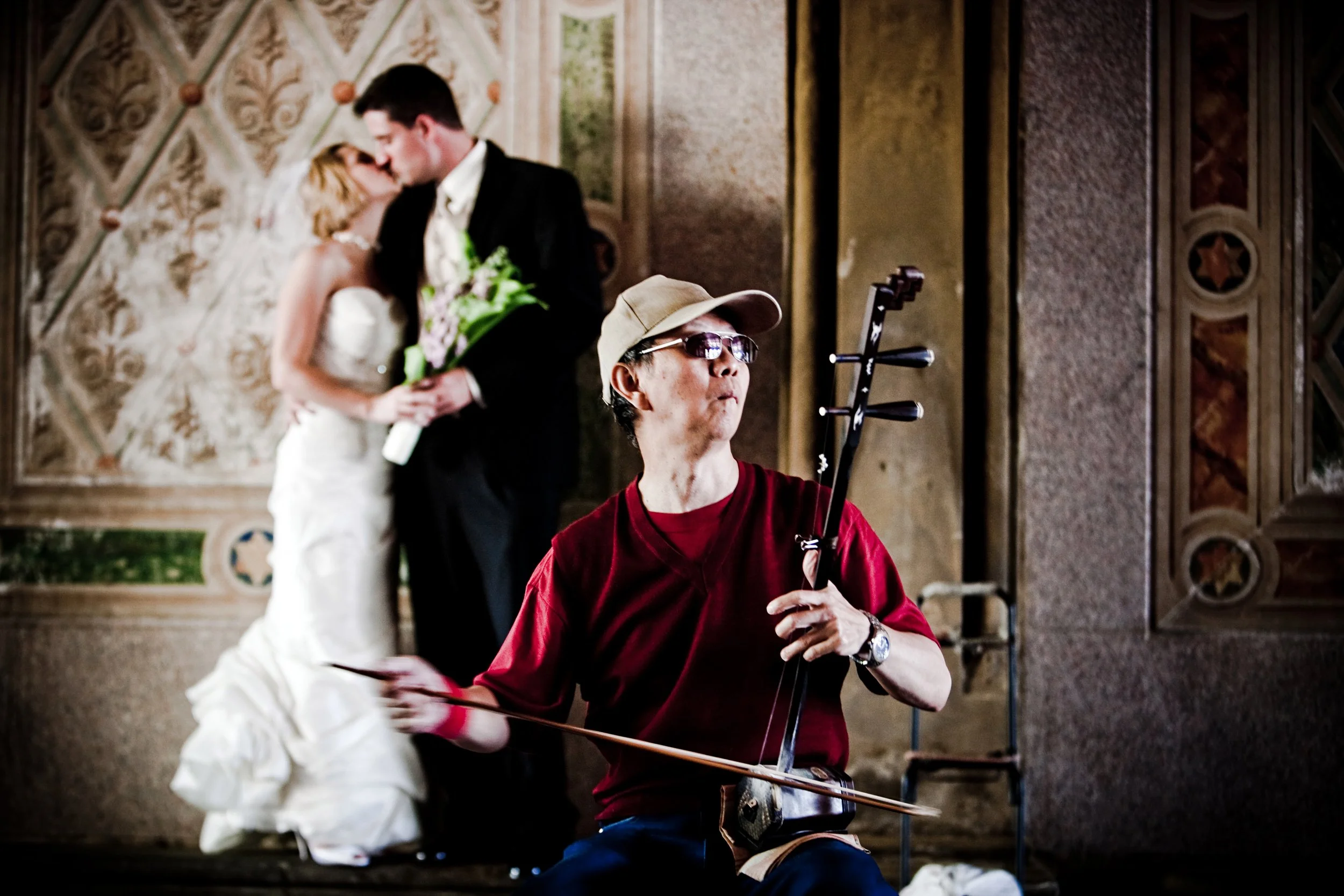 A musician wearing glasses, a beige cap, and a red shirt playing a traditional string instrument while a newlywed couple shares a kiss in the background.