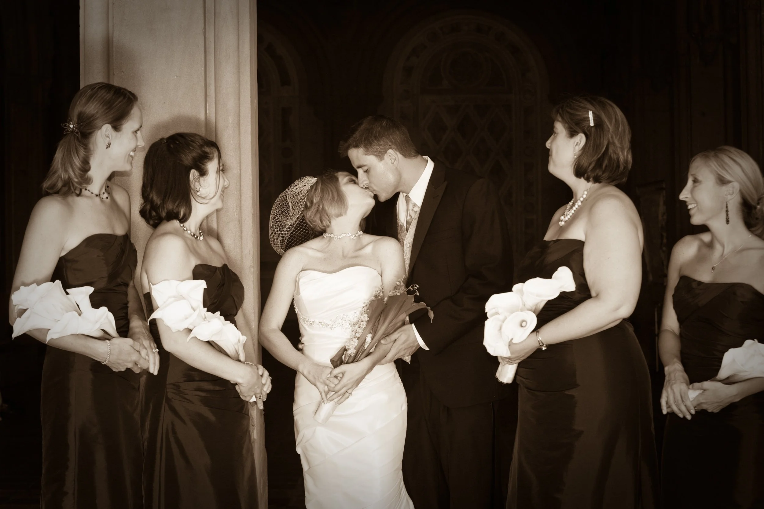 Sepia-toned photo of a bride and groom kissing, surrounded by bridesmaids holding calla lilies, during their wedding ceremony in a church.