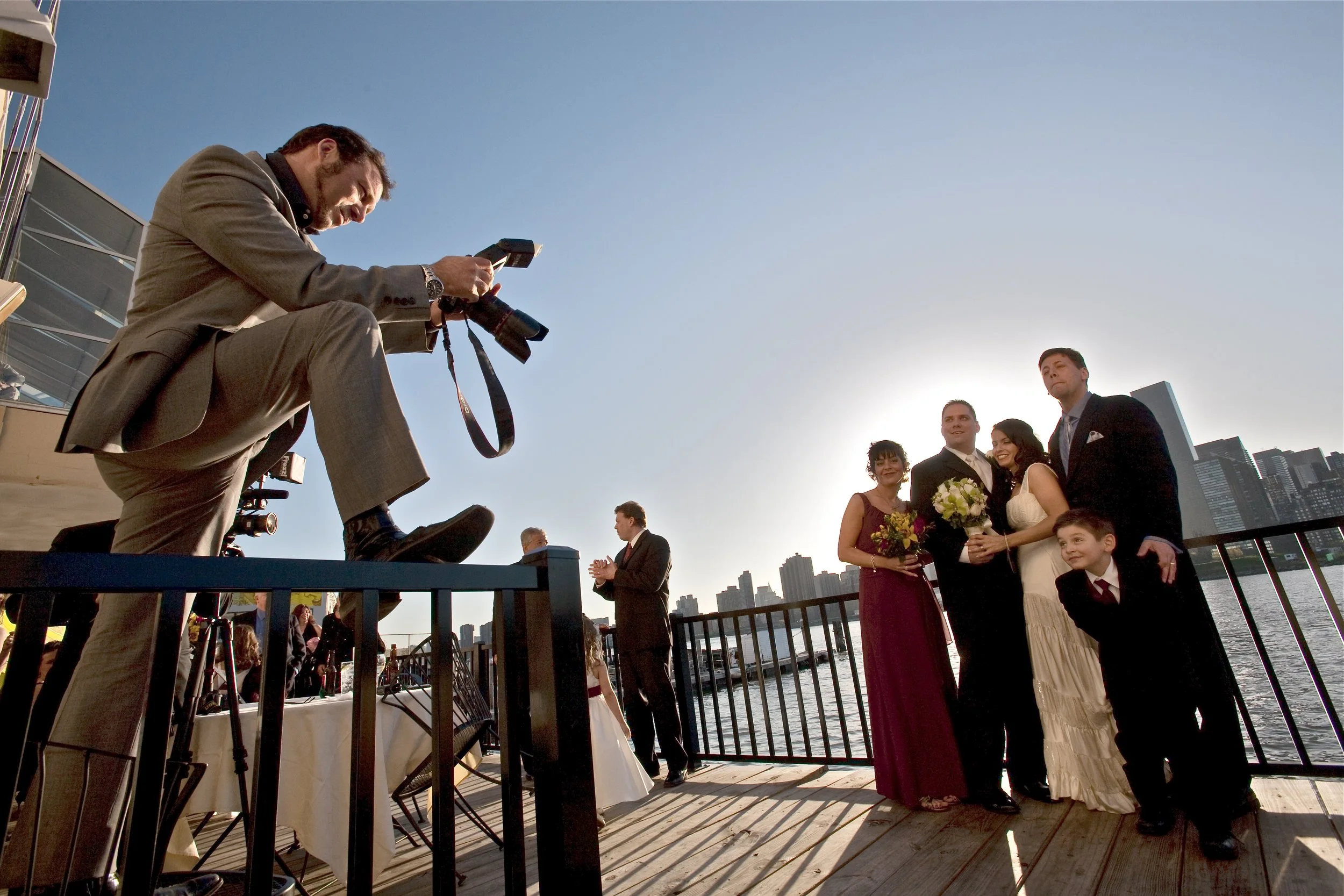 A wedding photoshoot on a riverfront dock during sunset with a group of people, photographer capturing the scene, and city skyline in the background.