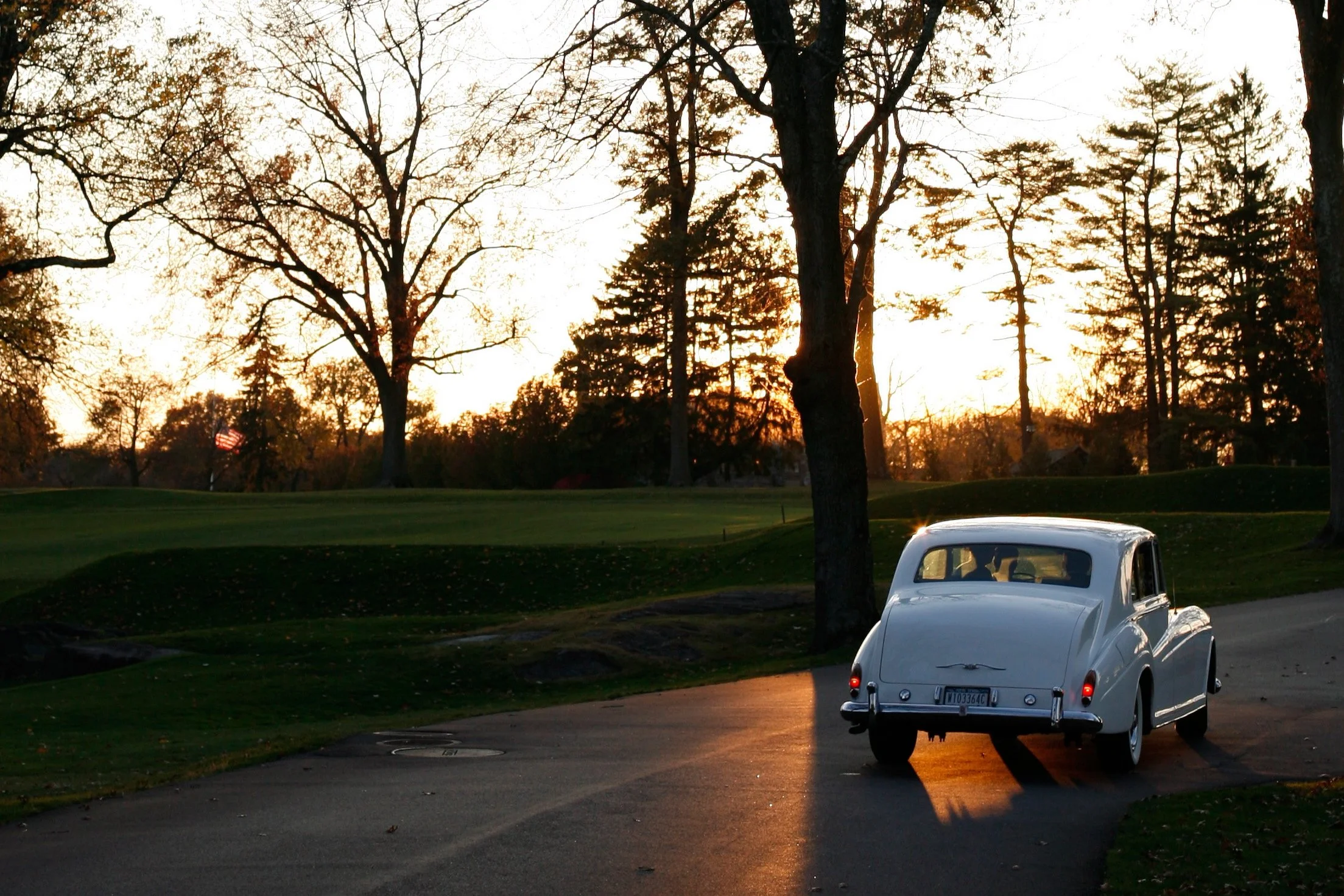 A white vintage car parked on a quiet road at sunset, with trees and a grassy area in the background.