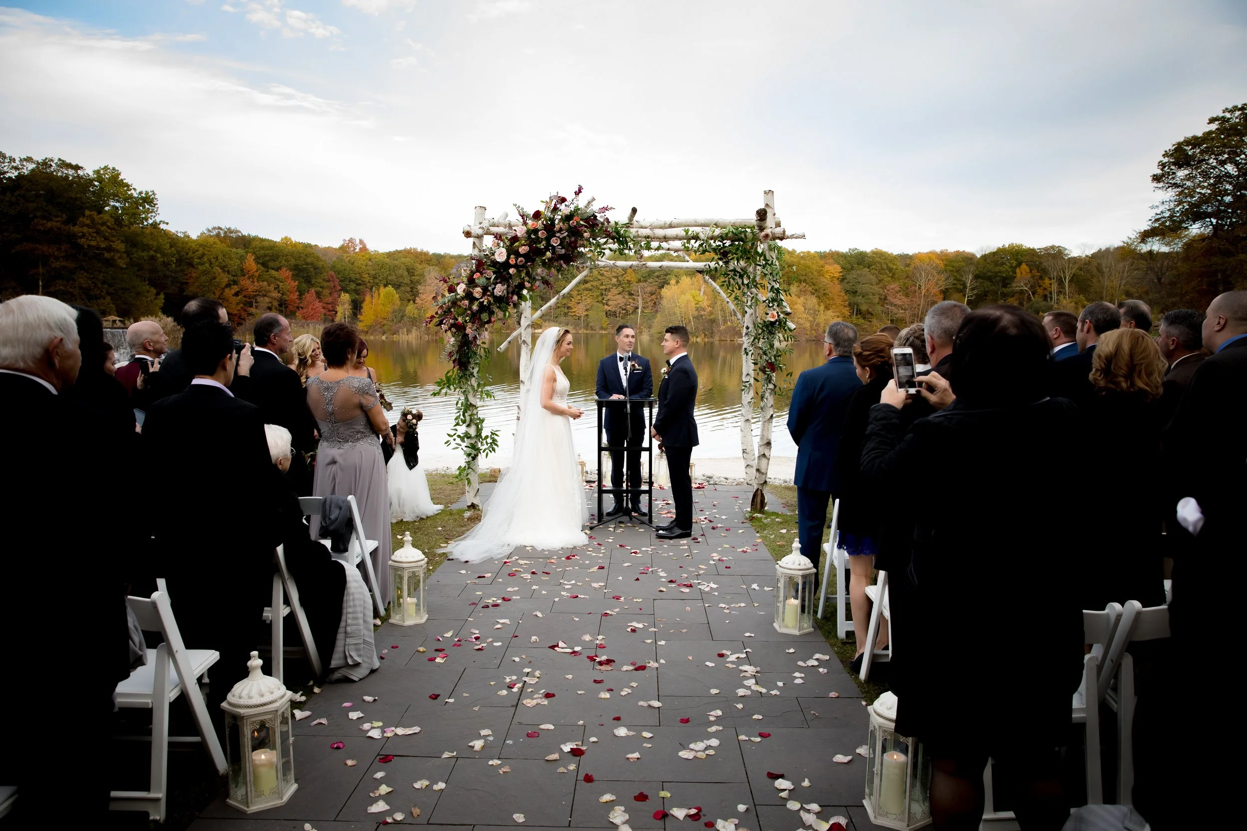 A wedding ceremony taking place outdoors near a lake with autumn-colored trees in the background. The bride and groom are standing under a floral arch while guests seated on either side watch and take photos. Rose petals and lanterns decorate the walkway.