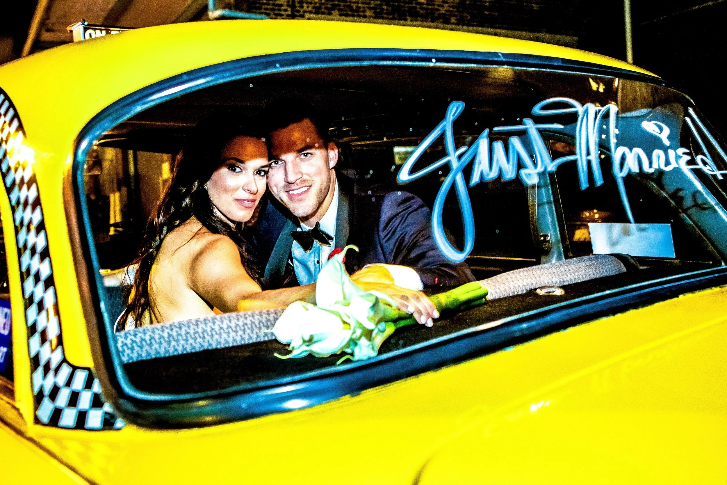 A newly married couple sitting in a yellow taxi cab, with the bride holding a bouquet of white calla lilies, both smiling and dressed in formal wedding attire, inside the car.