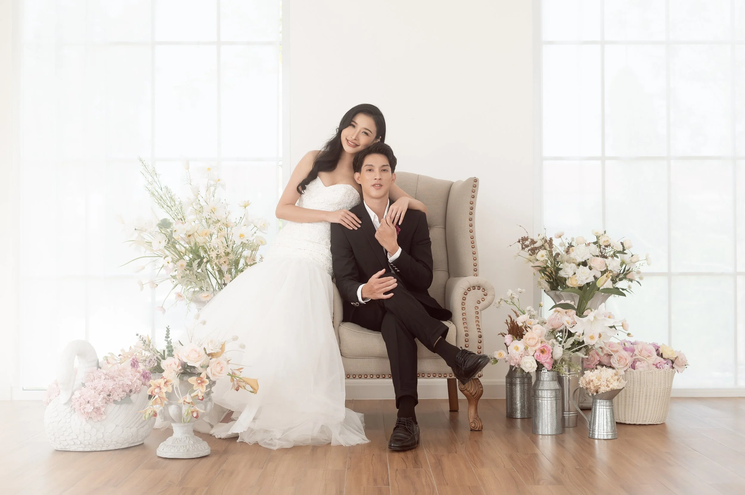 A newlywed couple dressed in wedding attire posing in a bright room with large windows and decorated with flowers.