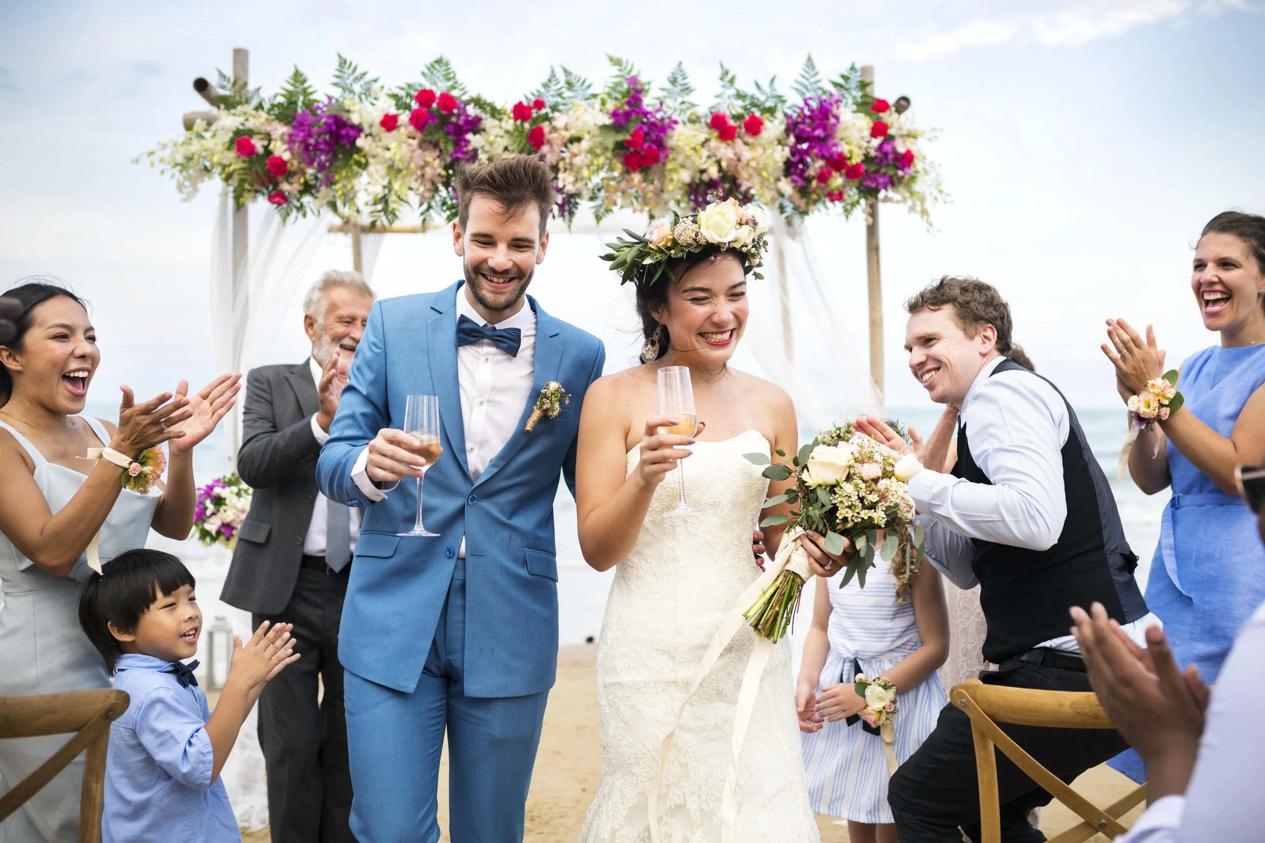 A wedding celebration on a beach with a bride and groom, surrounded by friends and family clapping and smiling, with a floral arch in the background.