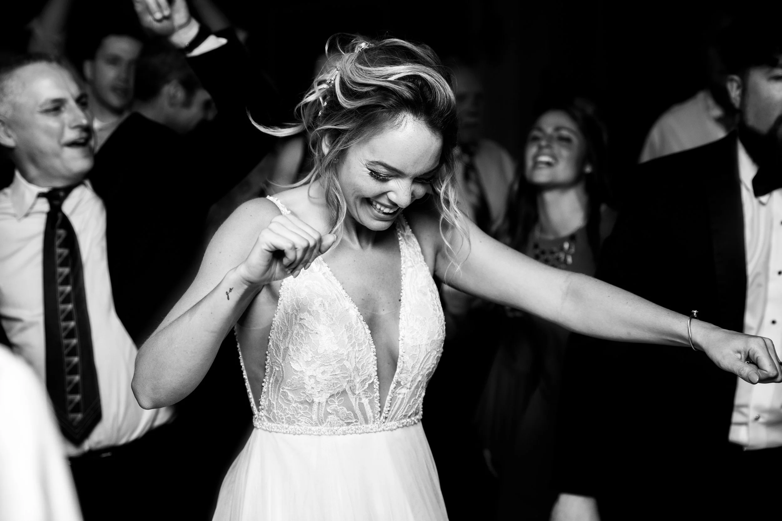 Black and white photo of a woman in a lace dress dancing and smiling at a celebration surrounded by people in formal attire.