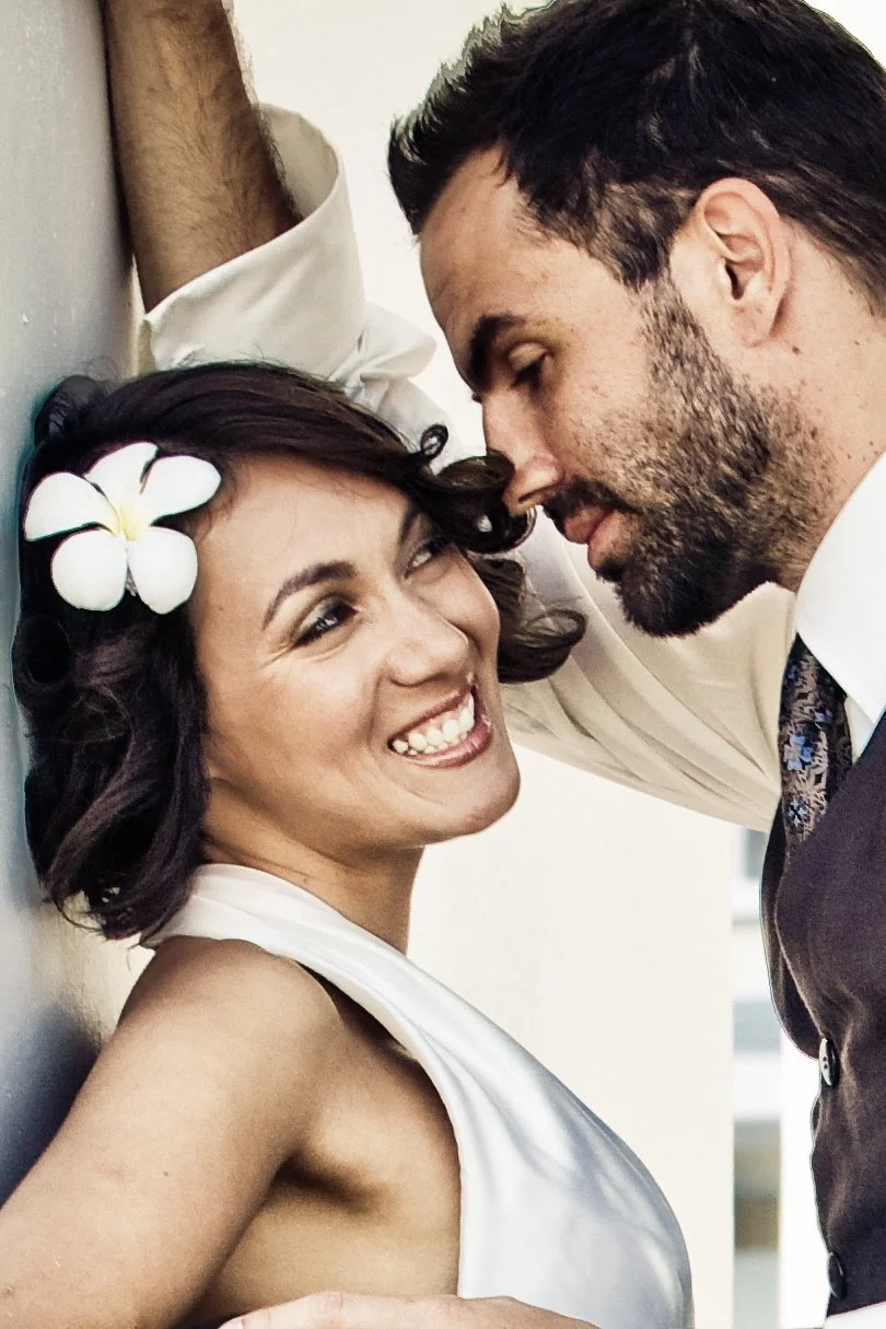 A woman with dark, curly hair and a white flower in her hair, smiling and looking at a man with dark hair and a beard, who is leaning close to her face.