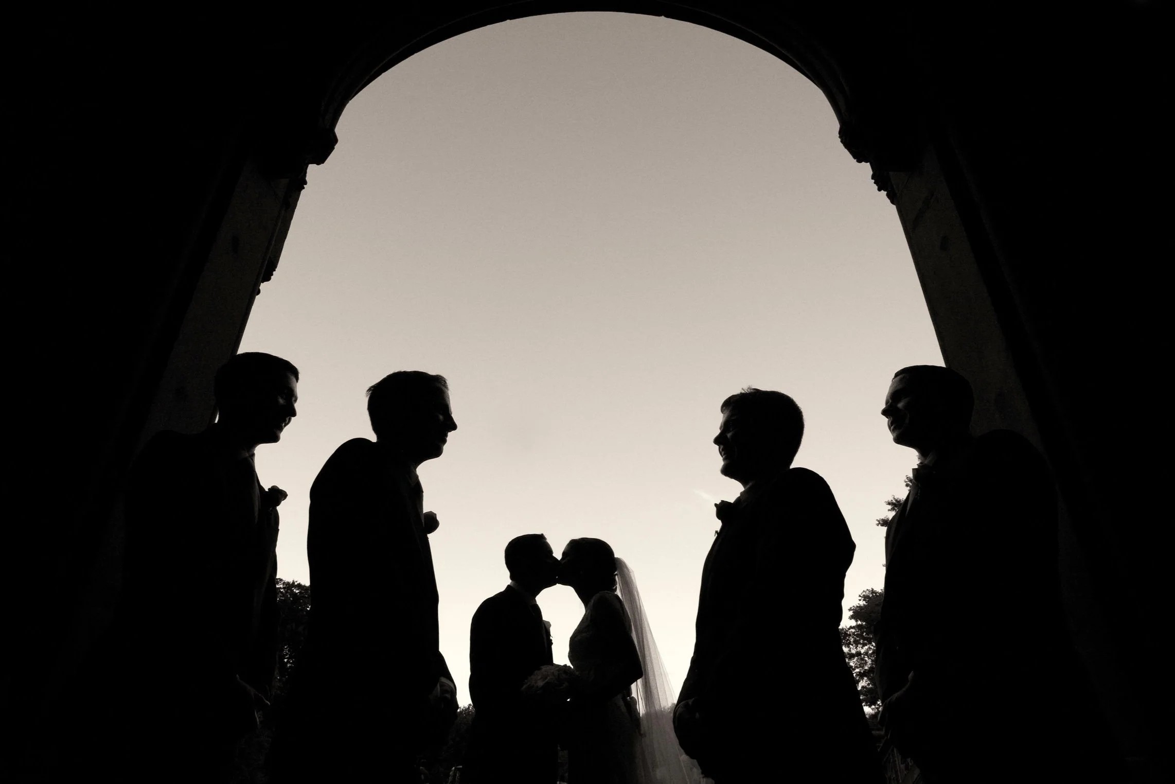 Silhouettes of a bride and groom kissing under an archway surrounded by four men, possibly groomsmen, against a dark sky.