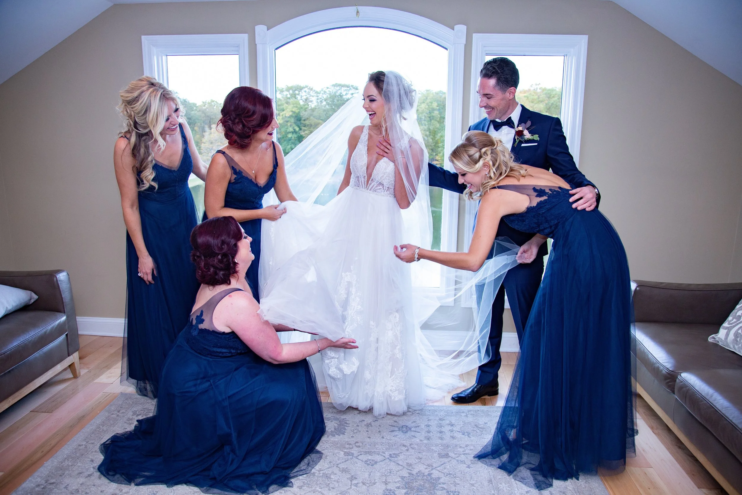 Bride in wedding dress surrounded by bridesmaids and groom in a room with large window