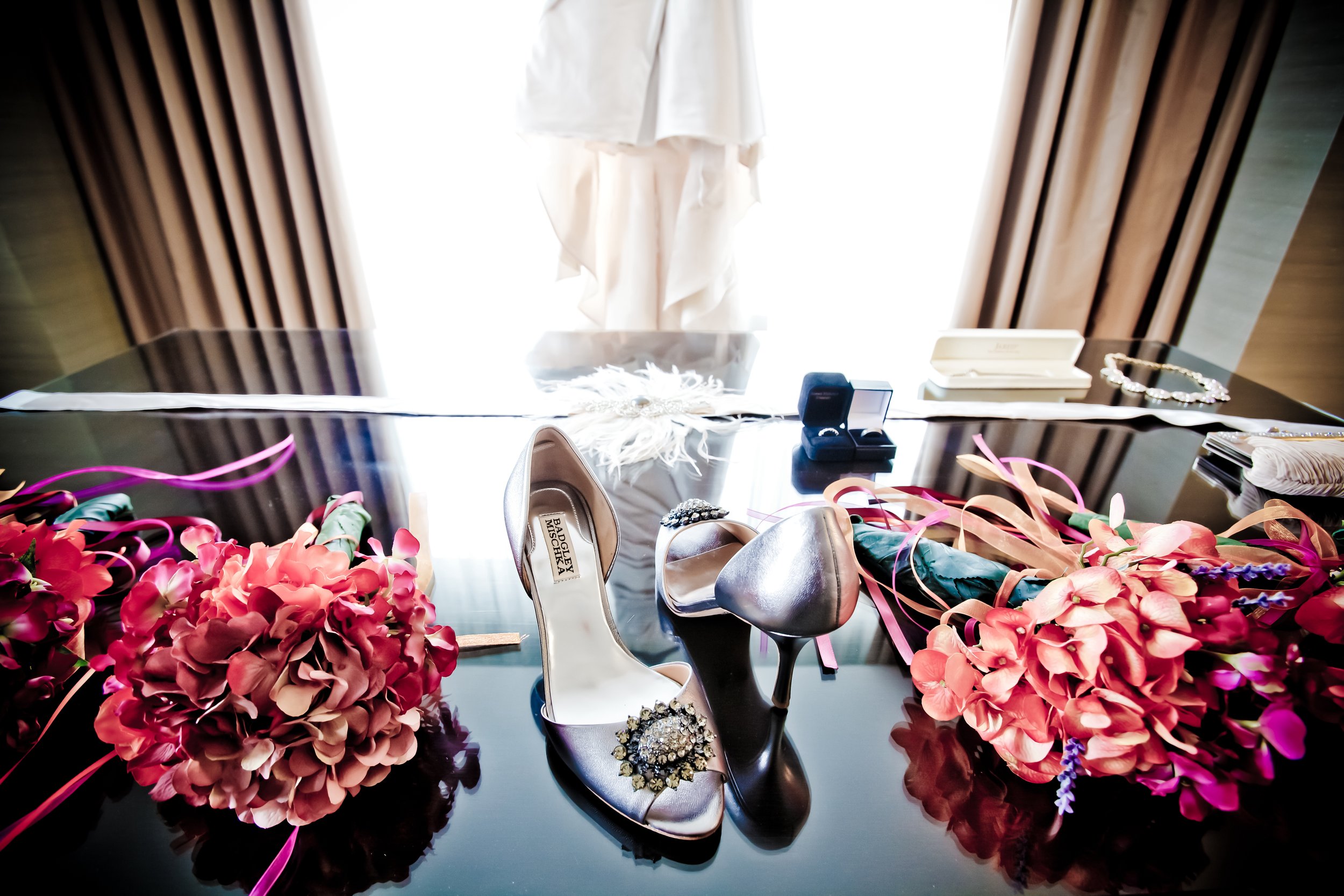 Wedding shoes, bouquets of pink hydrangeas, jewelry, and accessories on a black reflective surface in front of a window with beige curtains.