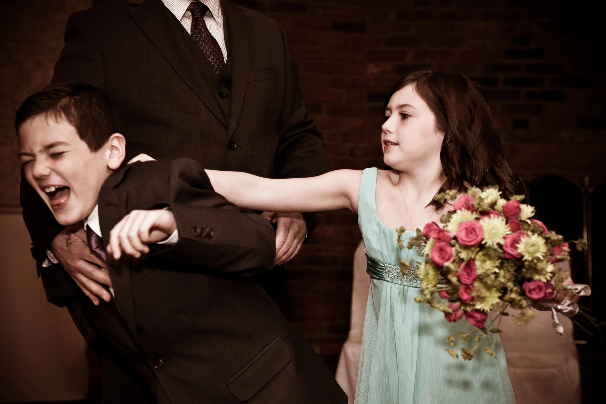 A girl in a light blue dress is grabbing a boy by the collar in a playful fight during a wedding reception. The girl is holding a bouquet of pink roses and yellow flowers. An adult man in a suit stands behind them.
