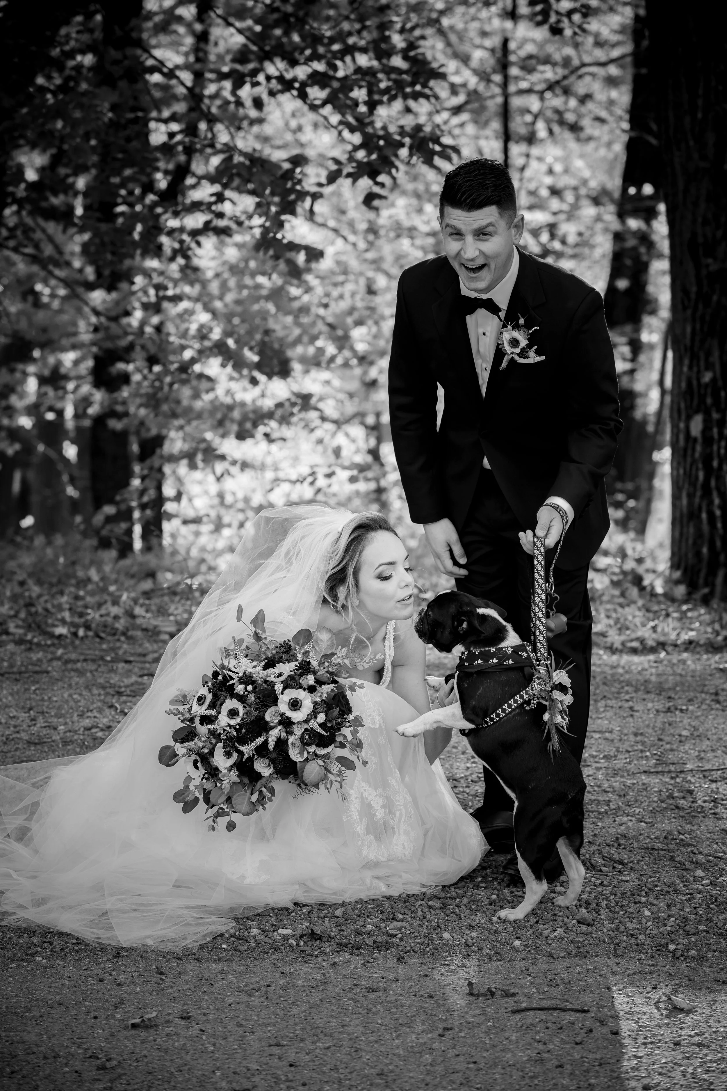 A black-and-white photograph of a bride and groom outdoors. The bride is squatting, leaning towards a dog, holding a bouquet, and wearing a wedding dress and veil. The groom, standing, is holding the dog’s leash, smiling at the dog. The background features trees and sunlight filtering through the leaves.