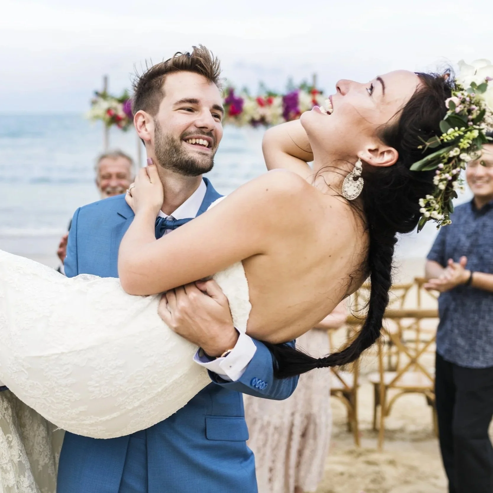 A newlywed couple dancing on the beach, with the groom holding the bride who is laughing and smiling, wearing a floral crown, during their wedding celebration.