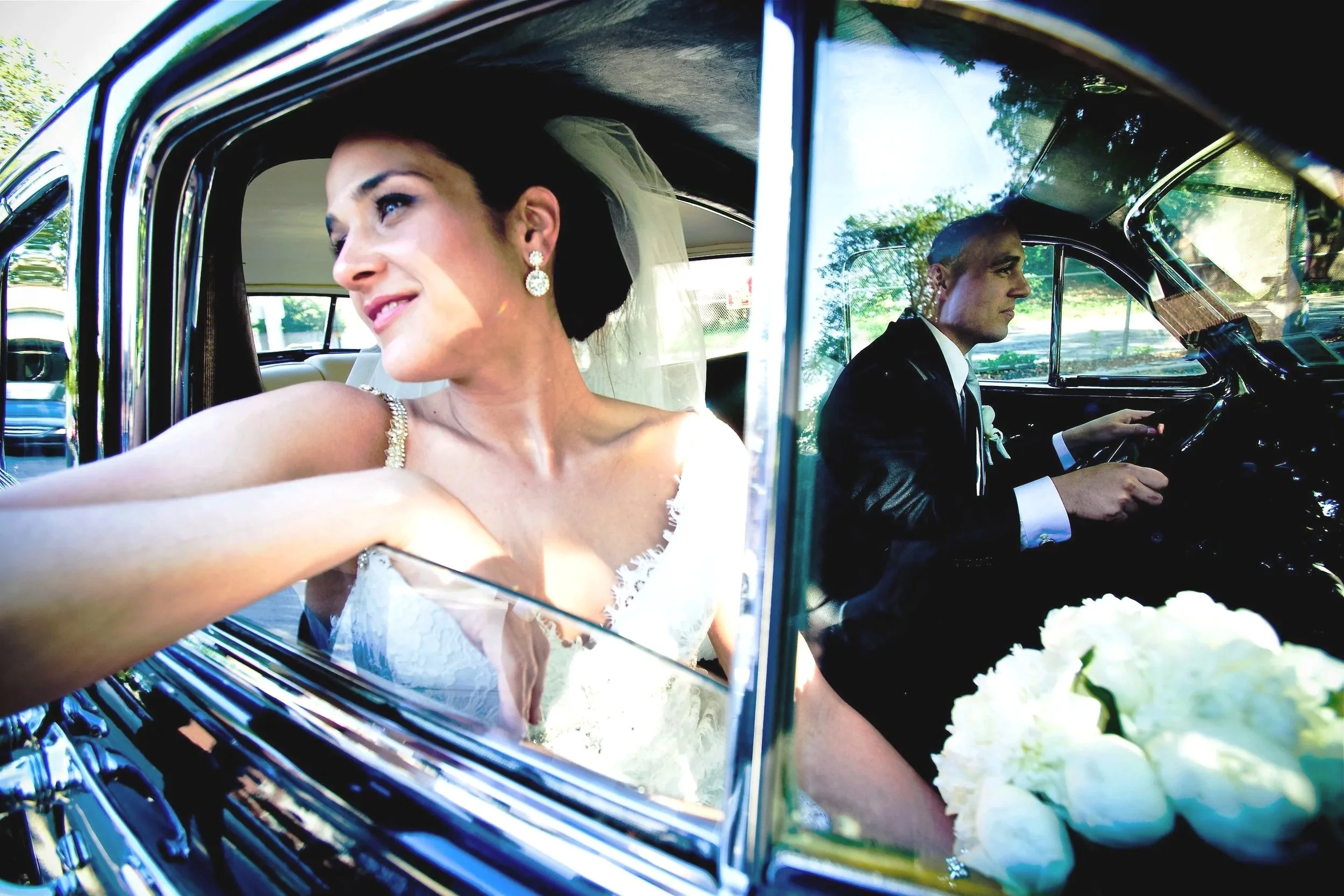 Bride in a white wedding dress sitting in the backseat of a vintage car with a groom driving, holding a bouquet of white flowers, with sunlight and trees visible outside.