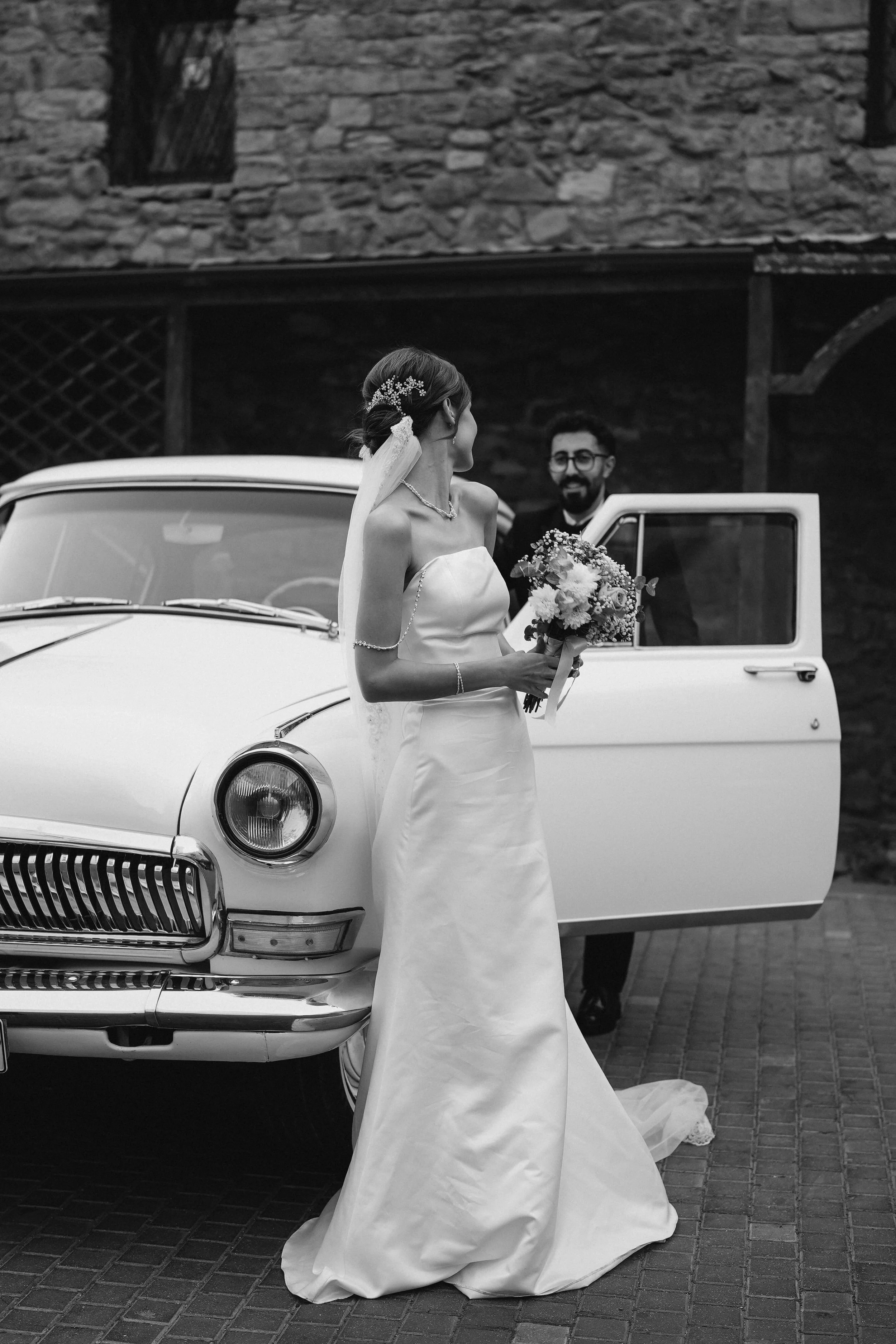 A bride in a wedding dress holding a bouquet, standing next to a vintage white car with an open door, smiling and looking at a man in a suit in the background.