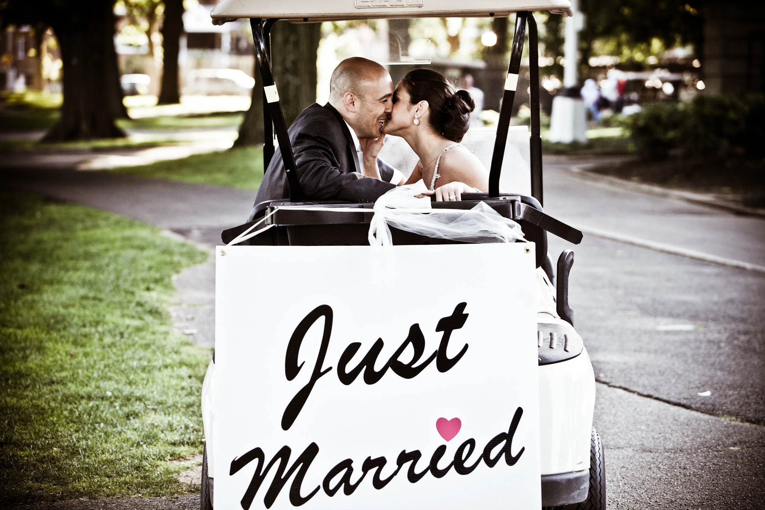 A newlywed couple in wedding attire sharing a kiss while sitting in a golf cart. A sign on the cart reads 'Just Married' with a small pink heart. The scene takes place outdoors on a sunny day with trees and a sidewalk in the background.