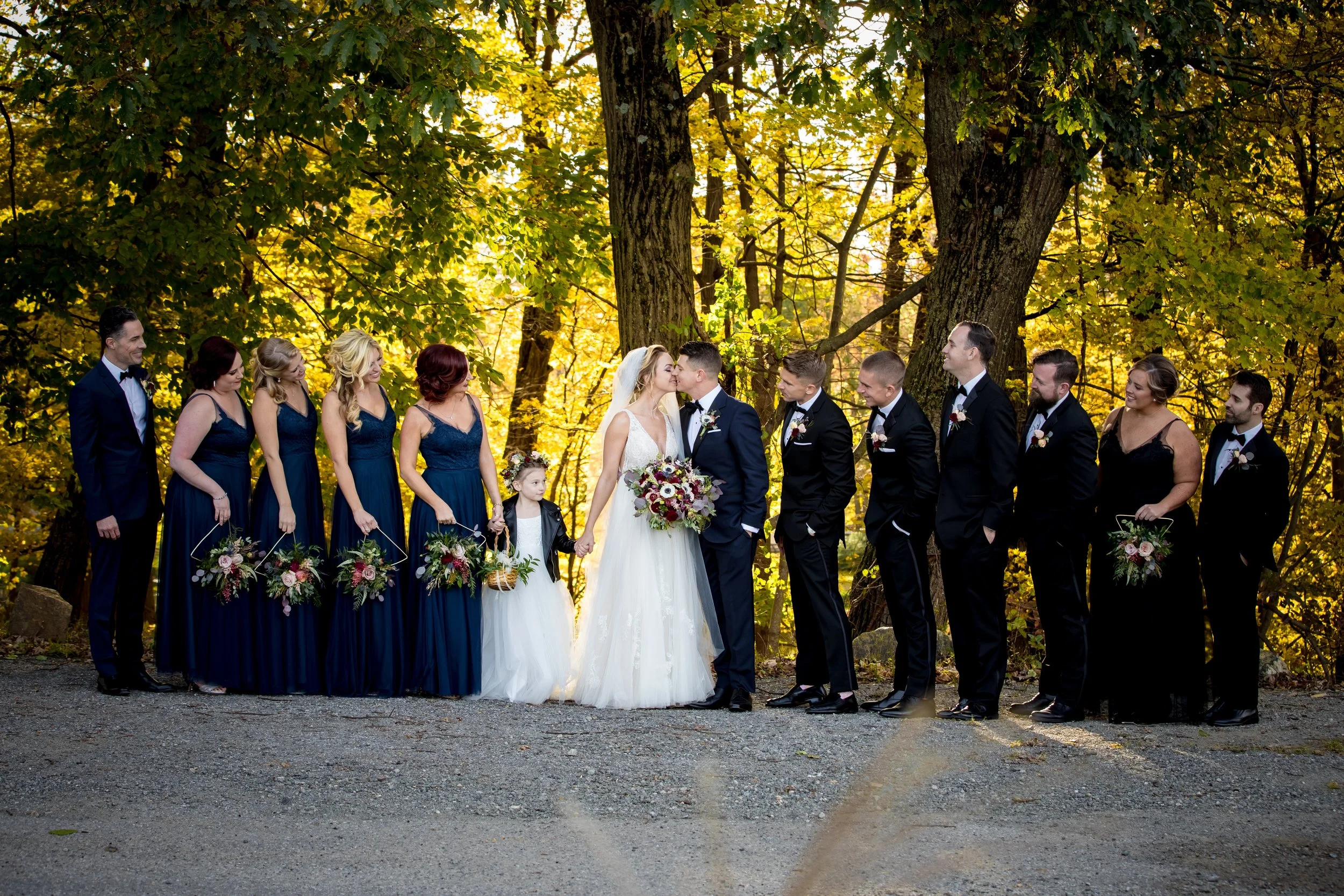 Wedding party standing outdoors in front of trees with yellow leaves; bride and groom in center kissing, holding bouquet, surrounded by bridesmaids holding flowers and groomsmen in tuxedos.