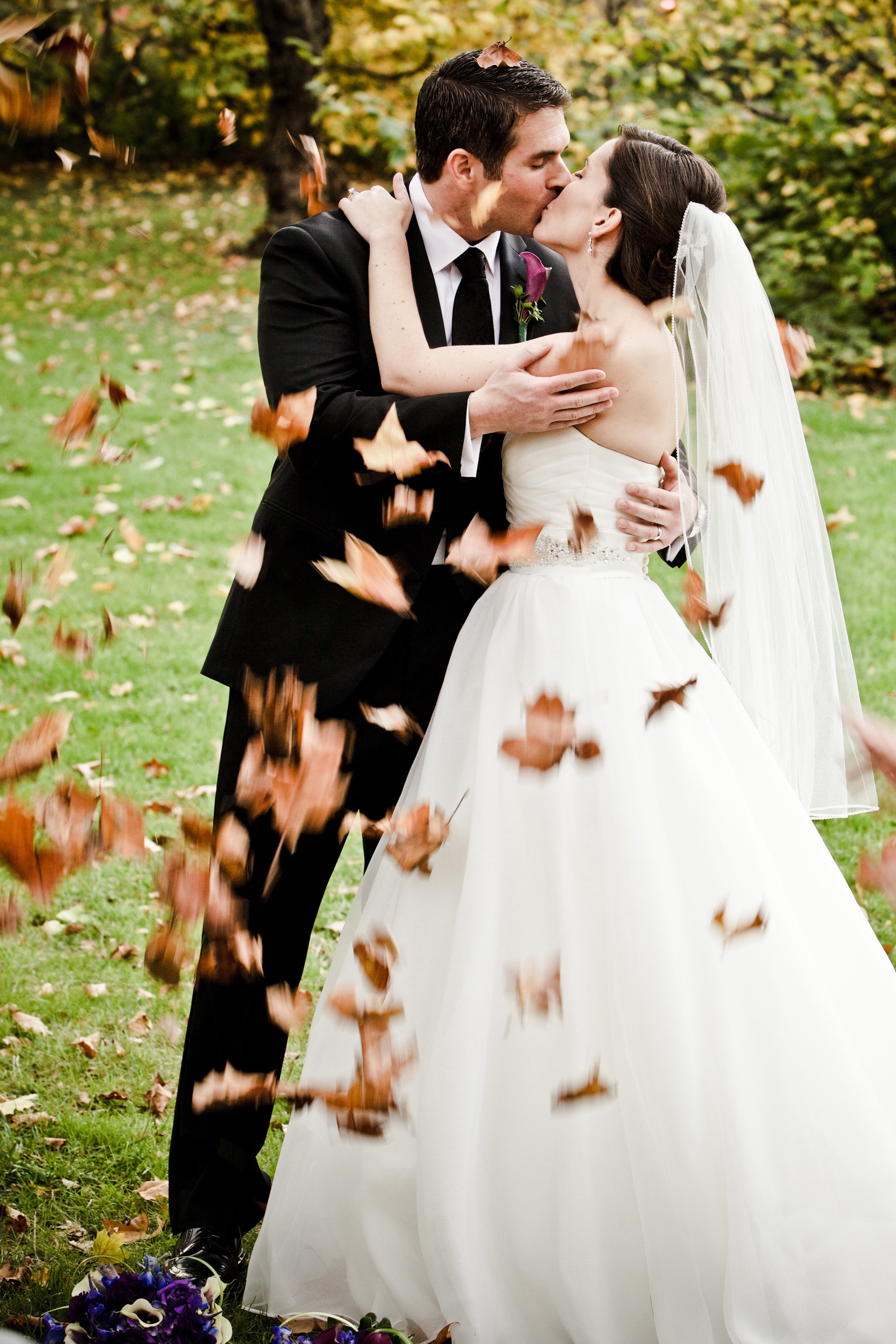 A bride and groom sharing a kiss outdoors during their wedding, surrounded by falling autumn leaves.