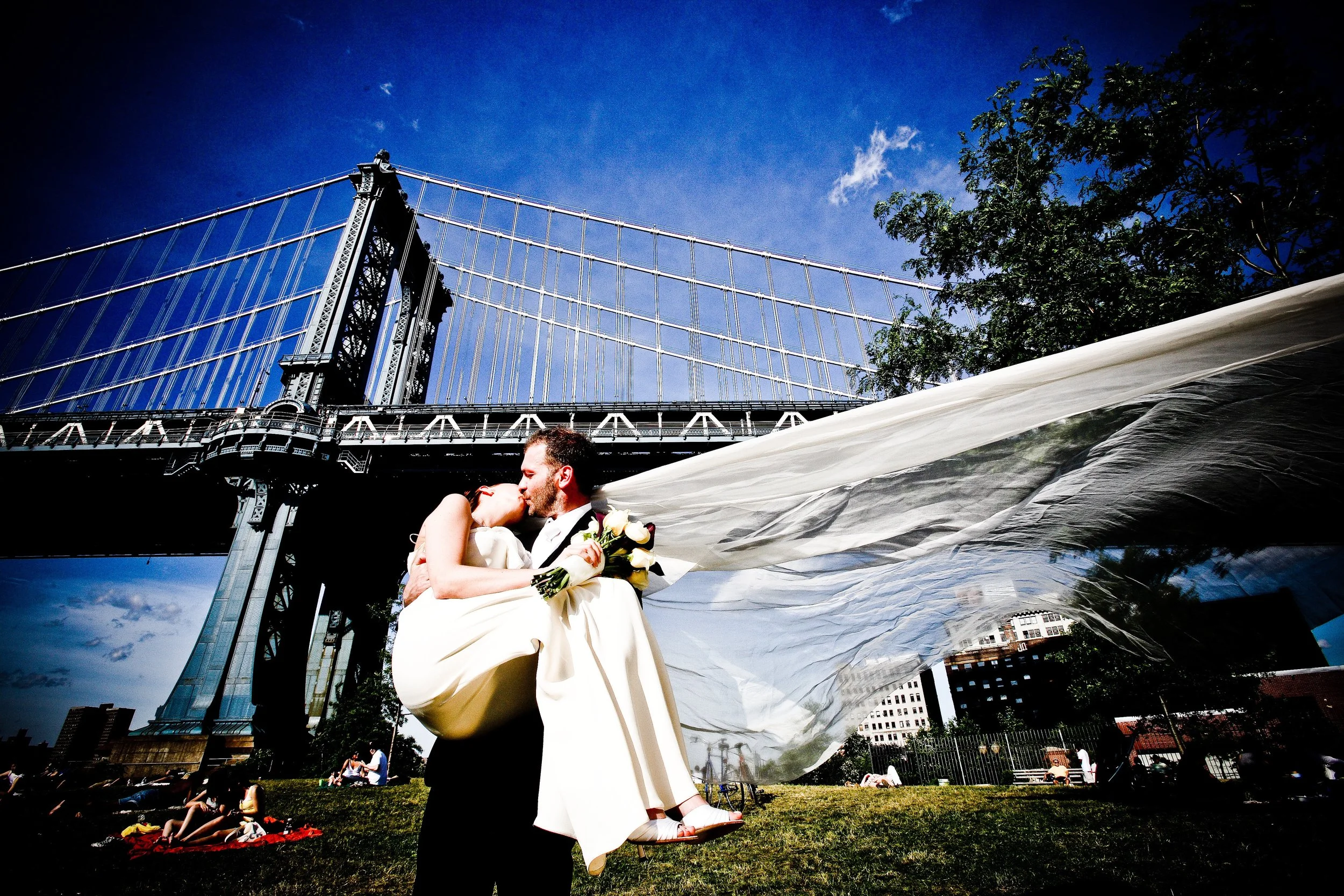 A groom holding a bride in a wedding pose under the Brooklyn Bridge on a sunny day.