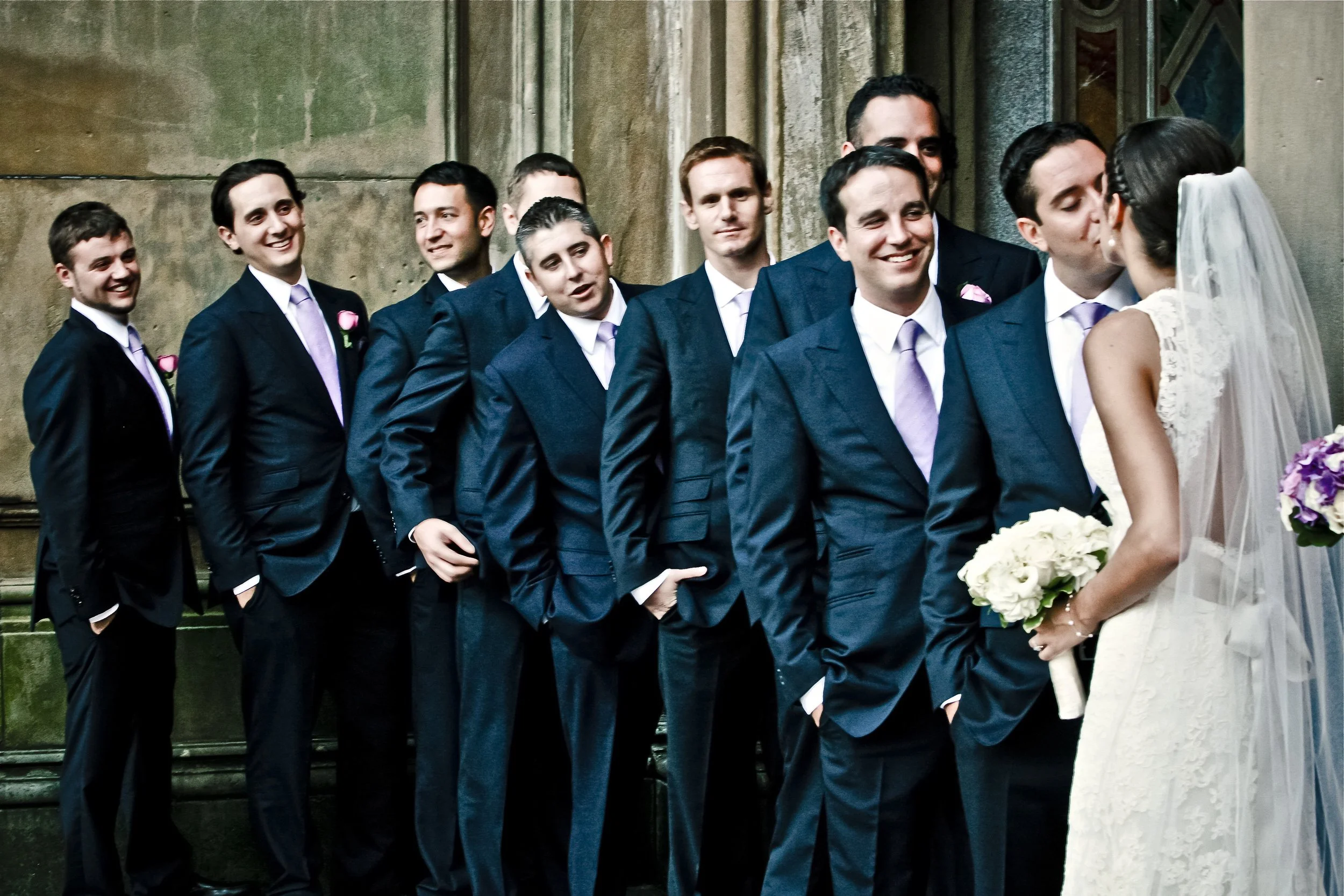 A bride and groom facing each other with the groom surrounded by his groomsmen standing in a line behind him, outdoors in front of a stone building.