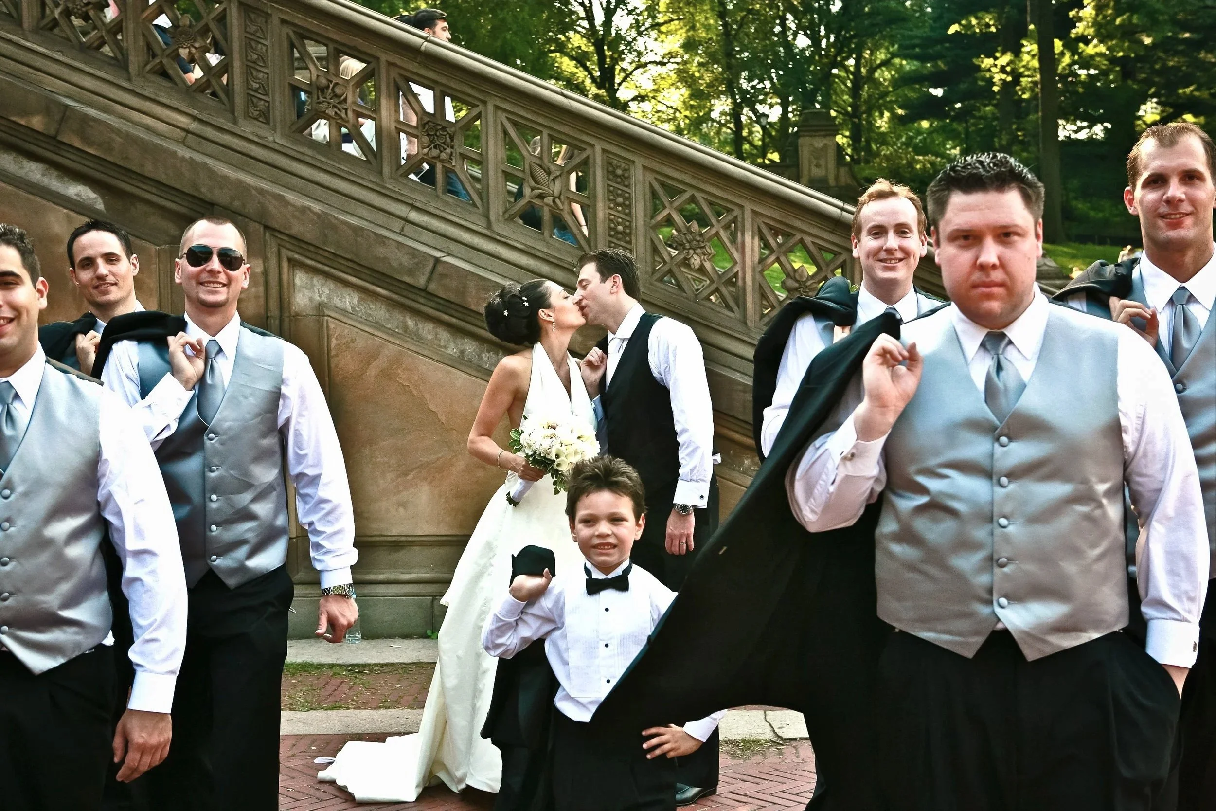 A wedding party outdoors with a bride and groom kissing in the background, surrounded by groomsmen and a young ring bearer in formal attire, near a stone staircase and lush green trees.