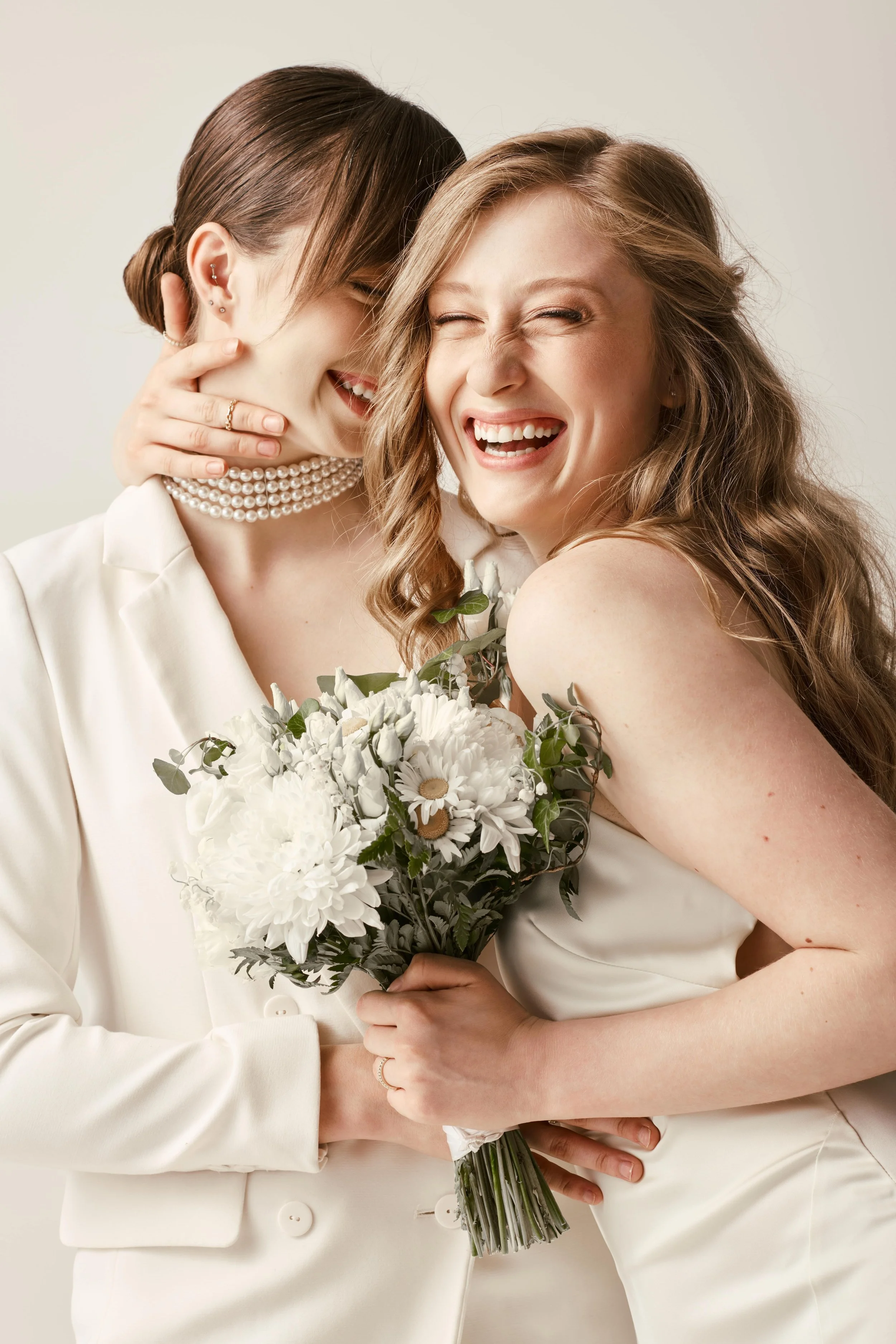 Two women in wedding attire sharing a joyful moment, one holding a bouquet of white flowers.