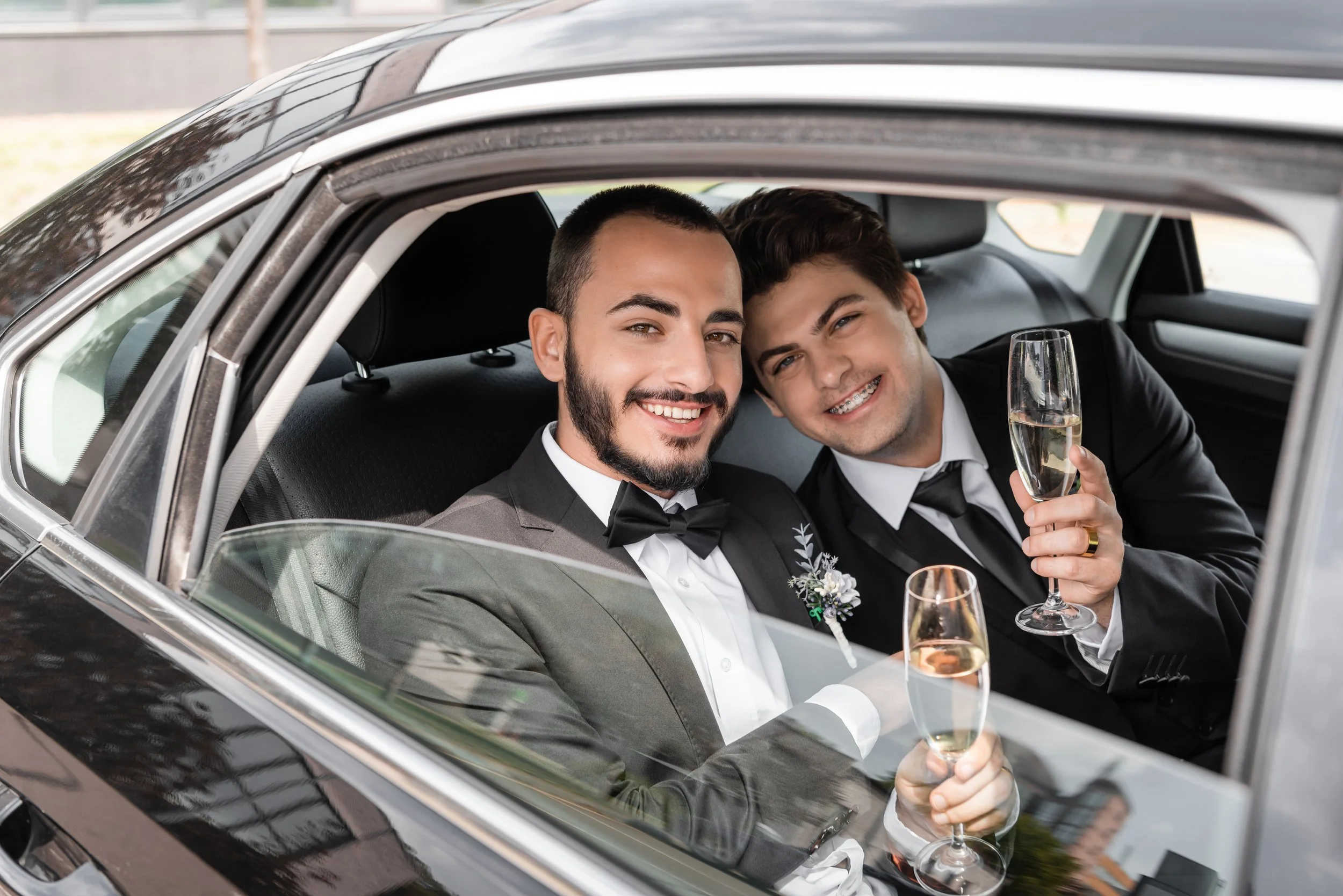 Two men in tuxedos celebrating with champagne inside a car.