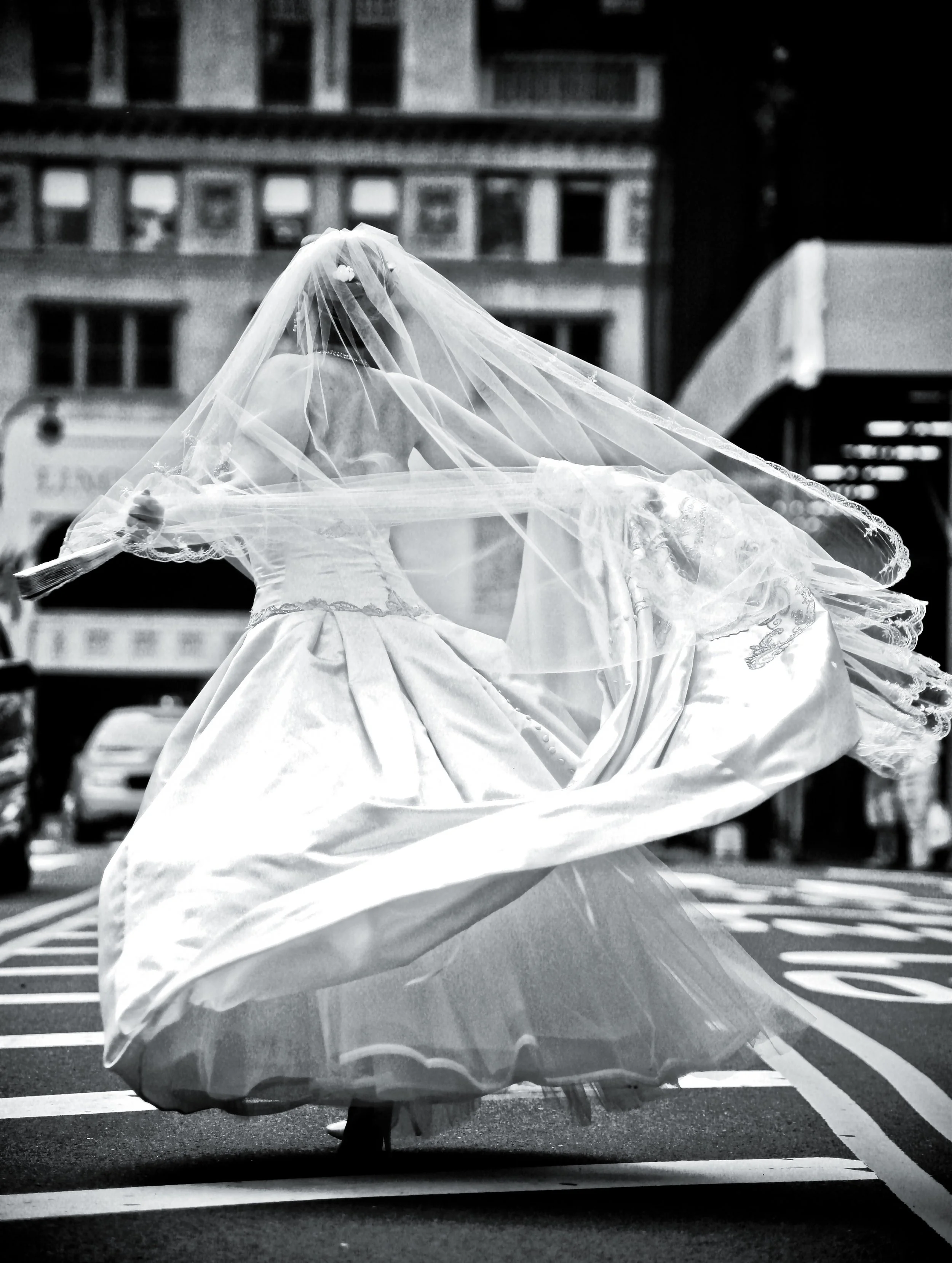 A bride in a wedding dress and veil stands on a crosswalk in an urban setting.