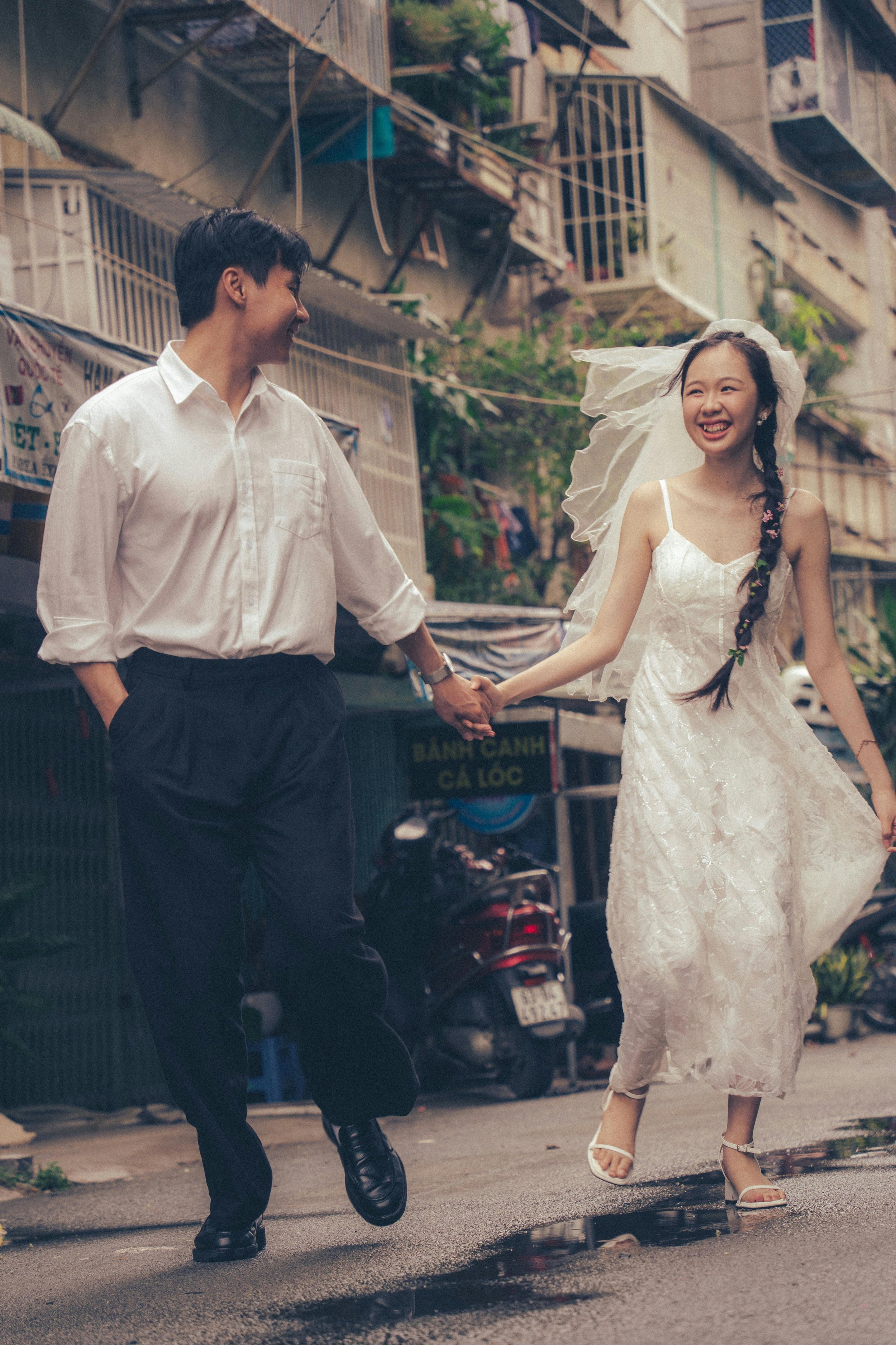A happy couple holding hands and smiling while walking on a wet city street, with the woman dressed in a white dress and veil, and the man in a white shirt and dark trousers.