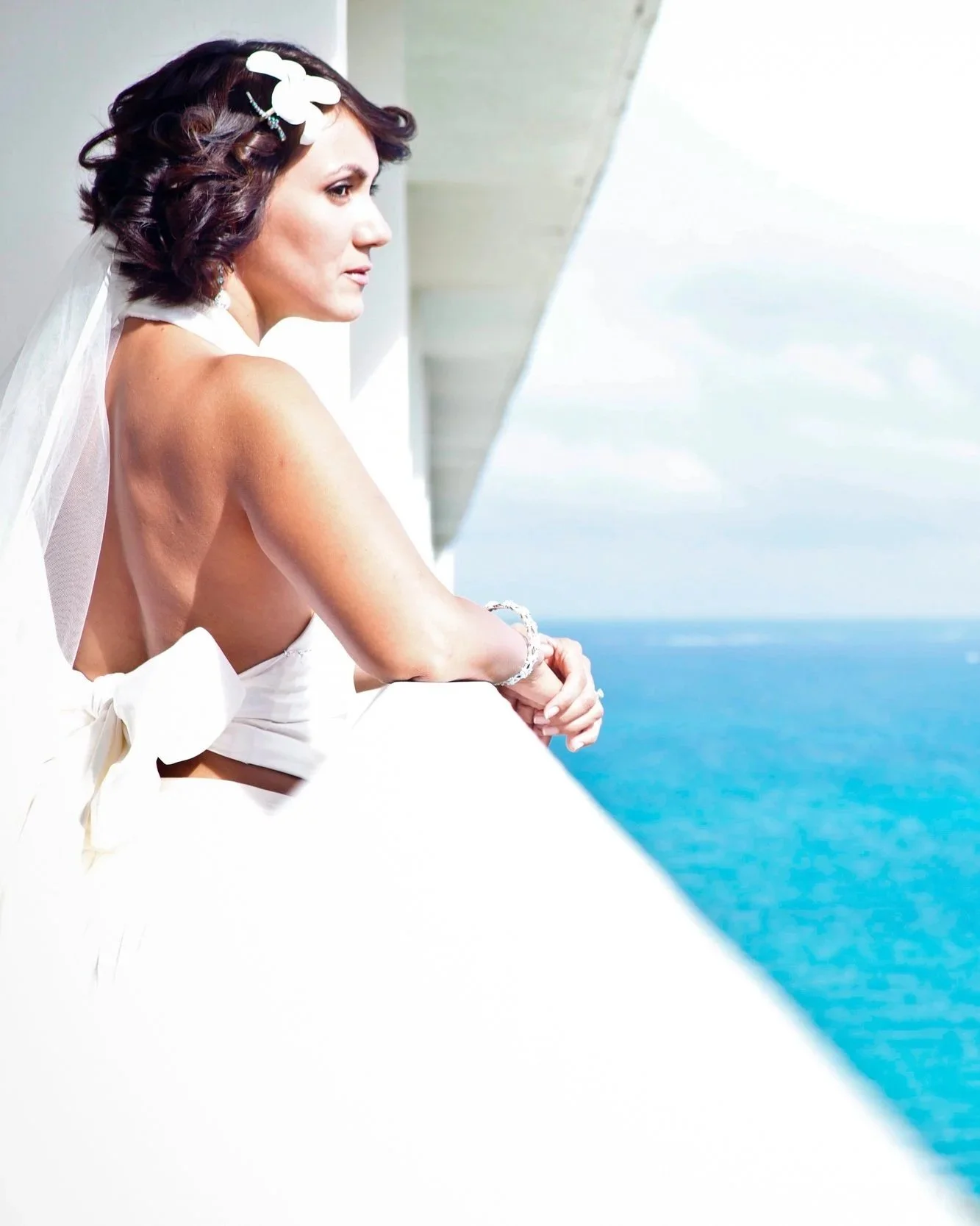 A woman in a bridal dress with a floral hair accessory and veil, leaning on a balcony railing, looking out over a blue ocean under a cloudy sky.