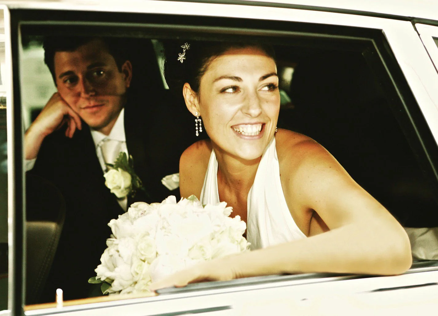 A bride and groom sitting in the back of a car, smiling. The bride is holding a bouquet of white flowers, wearing a white dress and earrings, with her hair styled up. The groom is wearing a black tuxedo with a white shirt and boutonniere, resting his chin on his hand.