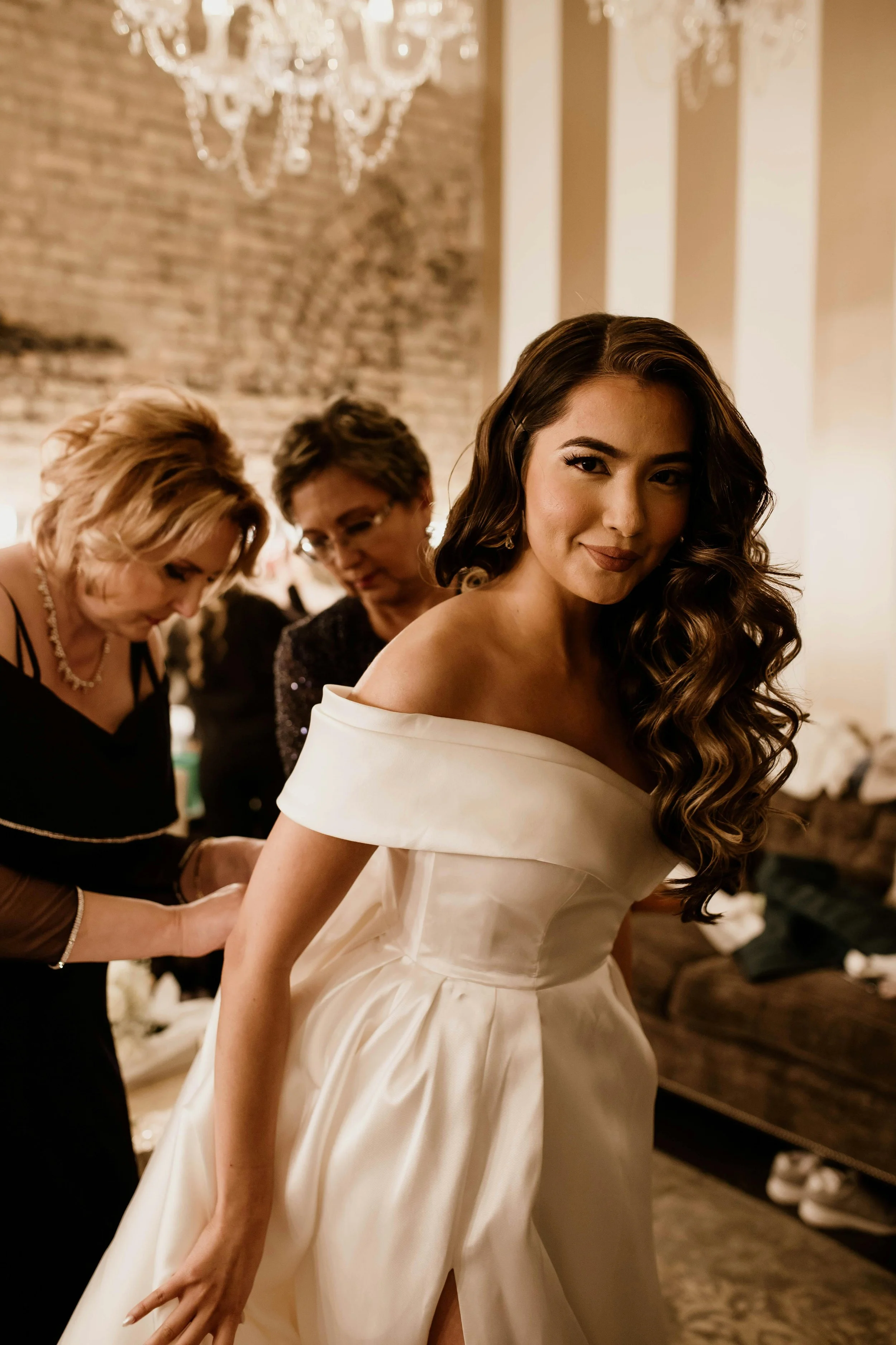 A young woman in a white wedding dress getting help from two women as she prepares for her wedding, in a room with exposed brick wall and chandelier.
