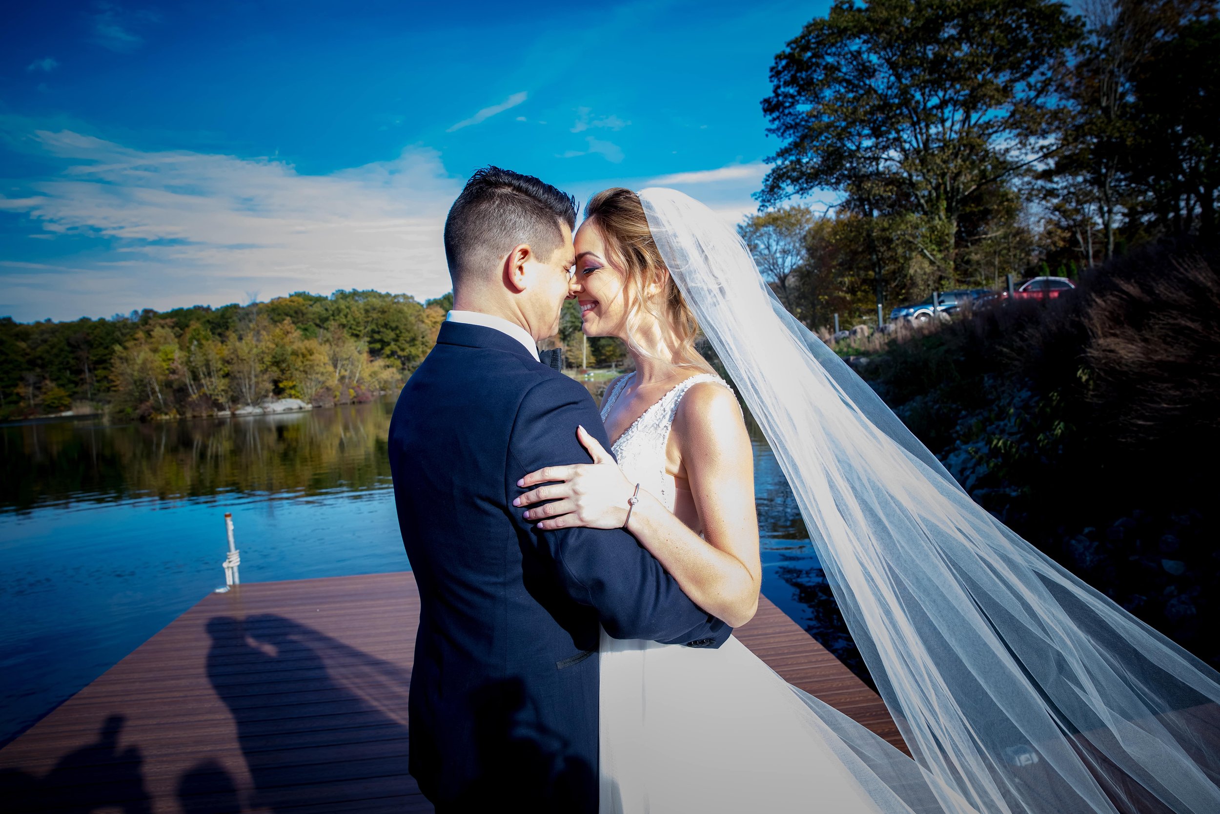 Bride and groom standing close by a lake, smiling, with trees and a blue sky in the background.