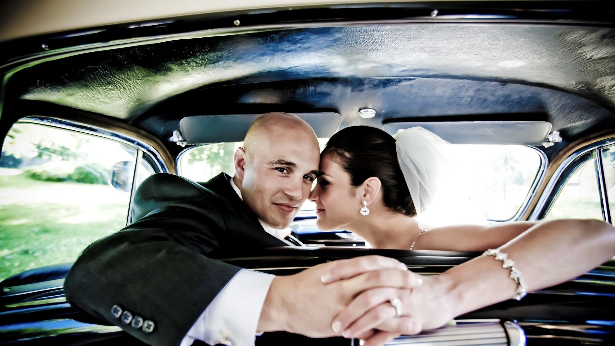 A newlywed couple in wedding attire sitting inside a car, leaning towards each other with their foreheads touching, smiling gently, with trees and a grassy area visible outside the windows.
