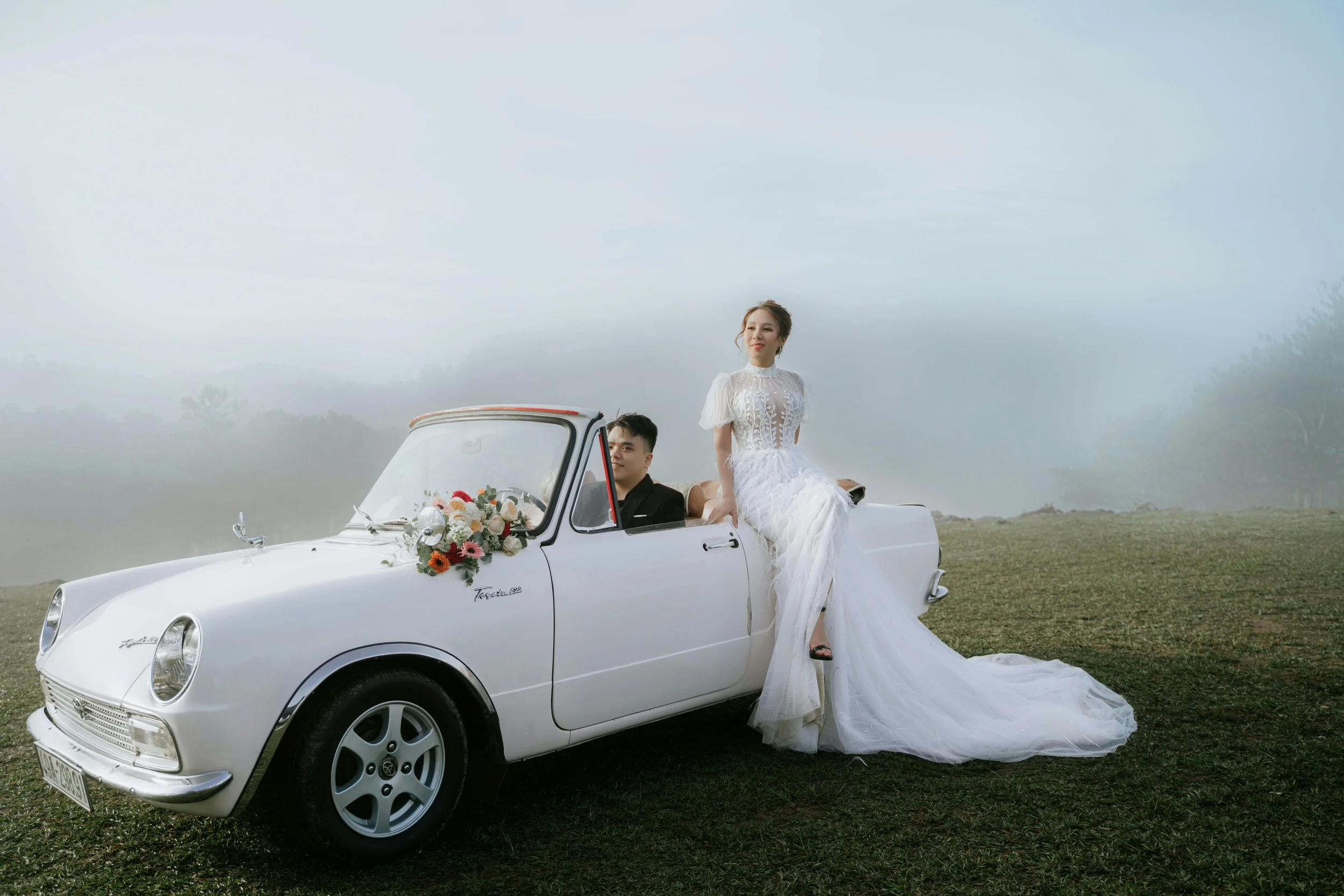 Bride in a white wedding dress sitting on the back of a vintage white convertible car decorated with flowers, with groom in a black suit sitting inside the car, outdoors on a grassy field with a foggy sky in the background.