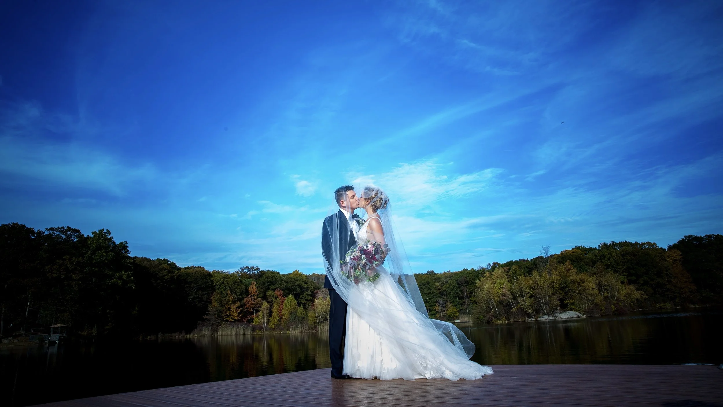 Bride and groom in wedding attire sharing a kiss on a dock near a lake, surrounded by trees with autumn foliage under a blue sky with wispy clouds.