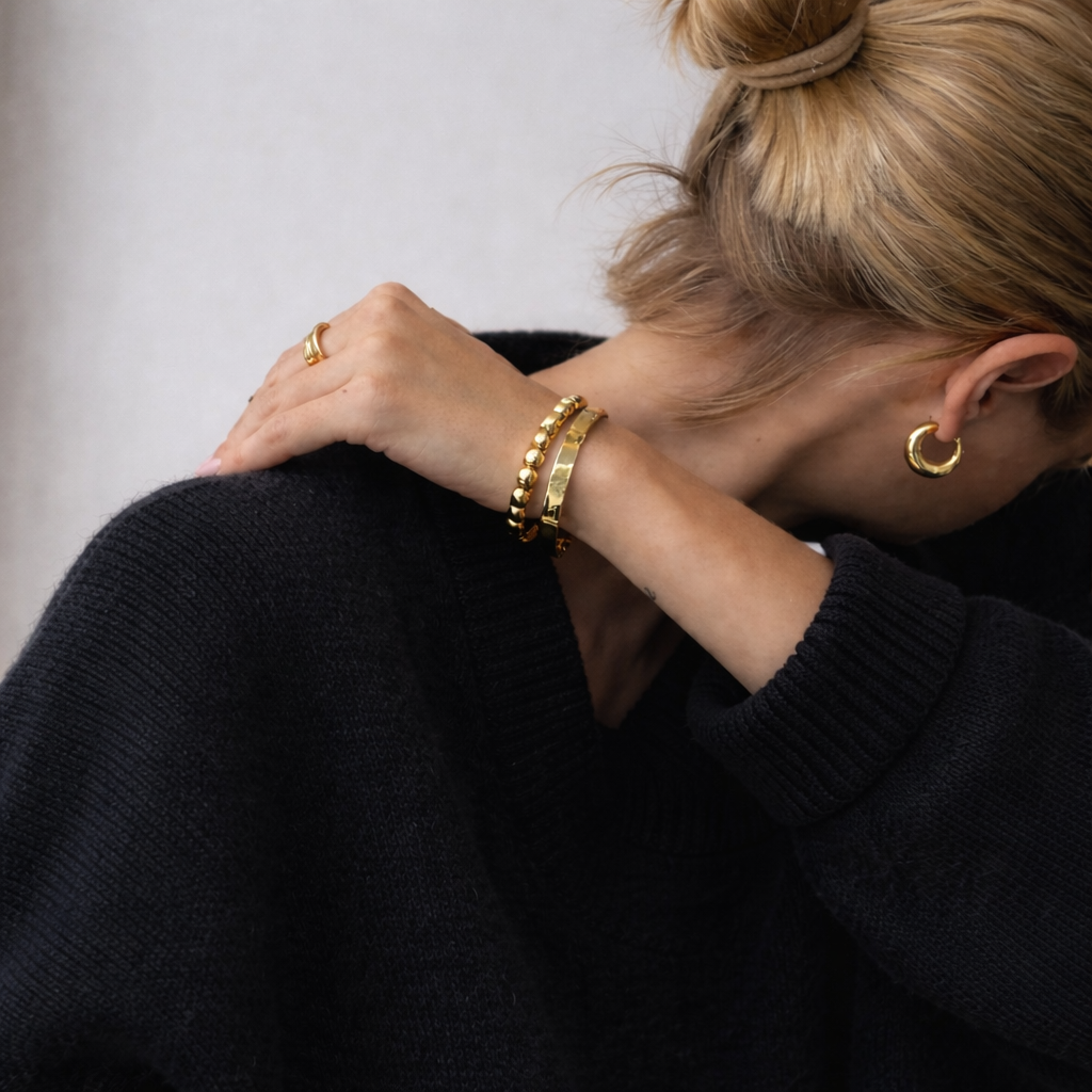 A woman with blonde hair wearing gold hoop earrings and gold bracelets, dressed in a black sweater, is sitting with her head down and her arms crossed over her shoulders.